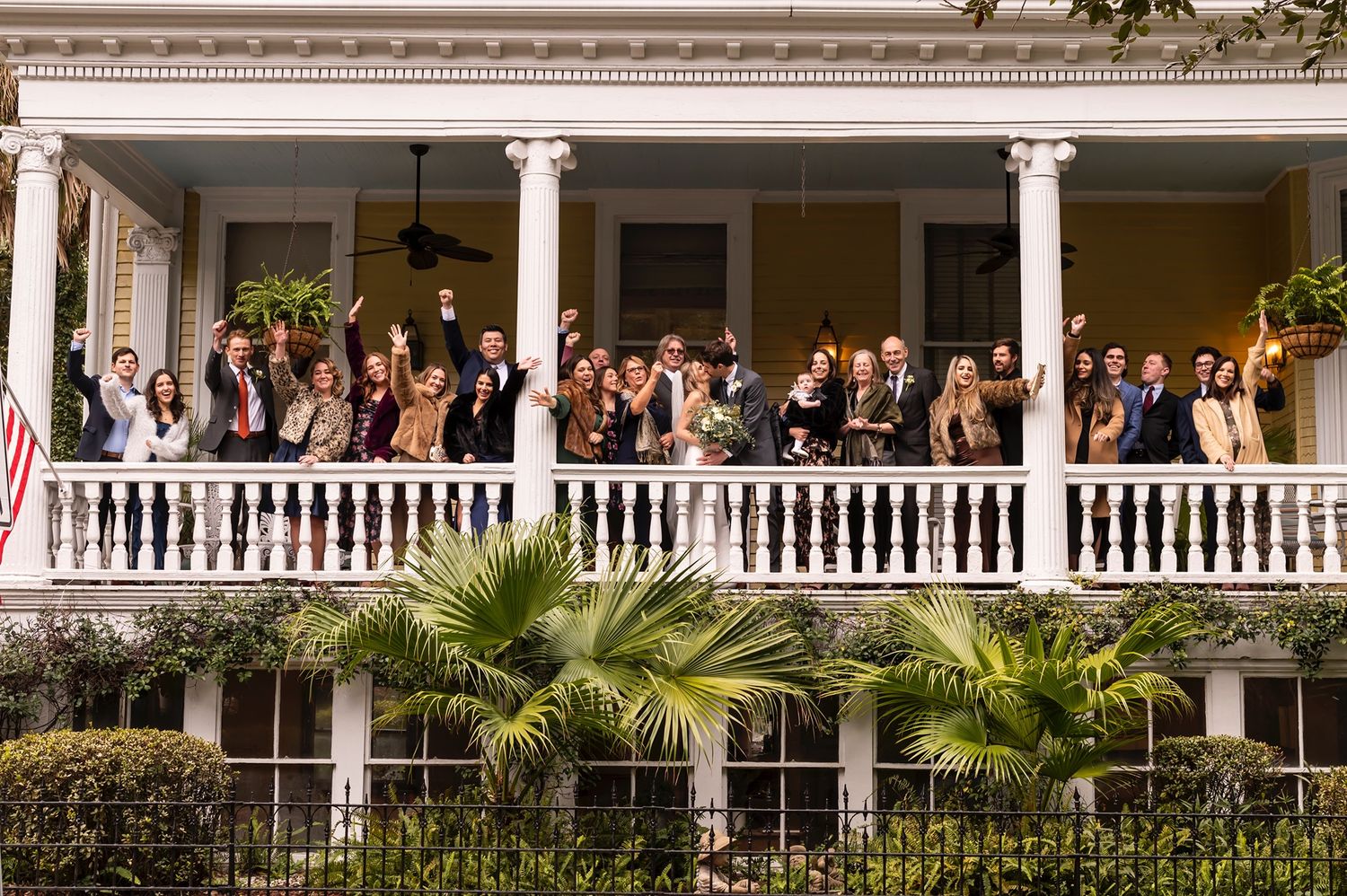 Large group of formally dressed guests standing along a white colonial-style veranda railing during an outdoor event.