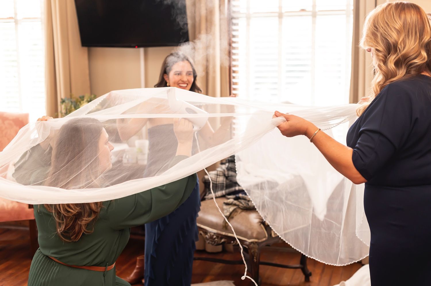 Three people hold up white tulle fabric in a living room setting while working together on a project.