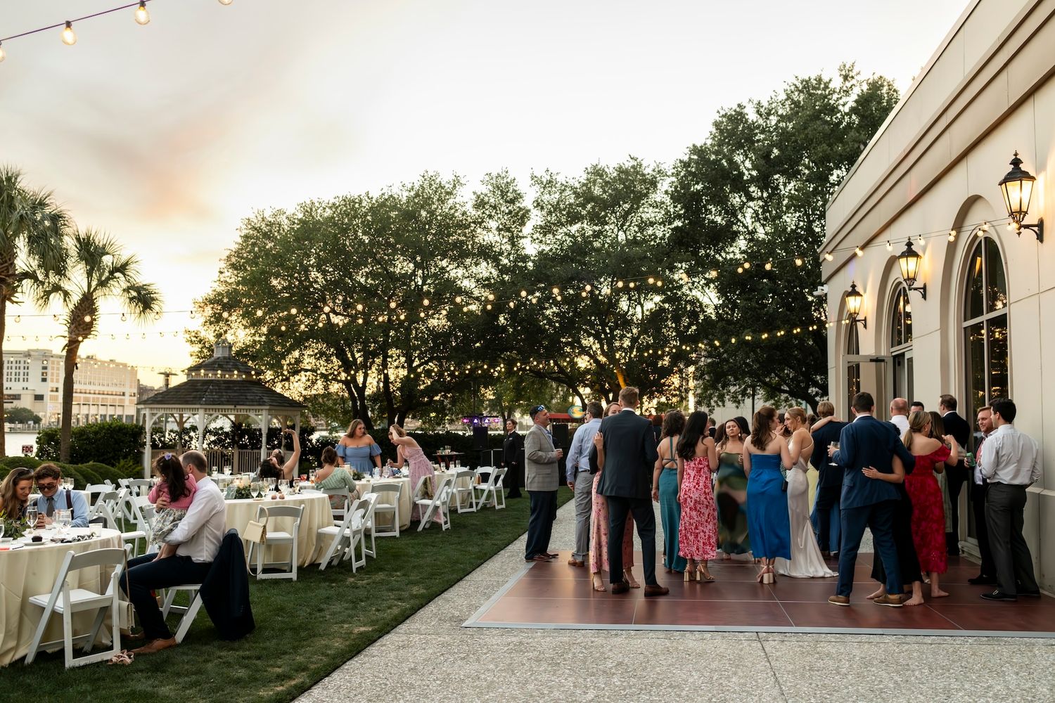 Outdoor wedding reception at dusk with guests dancing and dining on a patio adorned with string lights and lush trees.