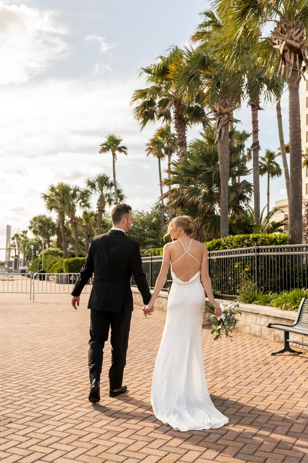 Bride and groom walk hand-in-hand along a brick path lined with palm trees, bride wearing a backless white gown.
