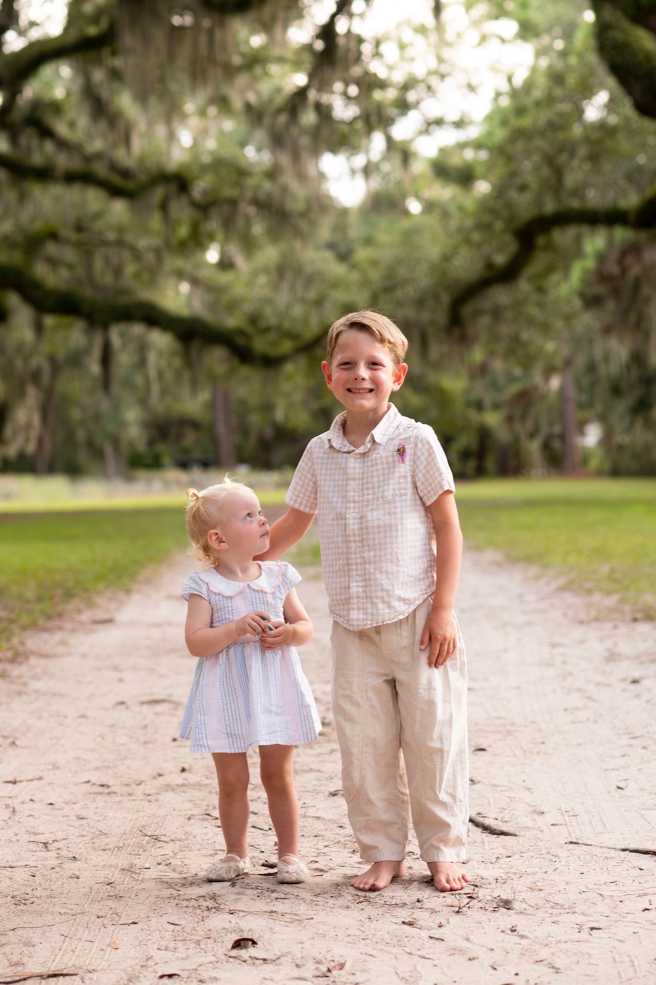 Two young children, a boy and girl, pose on a sandy path lined with moss-covered oak trees in a park.