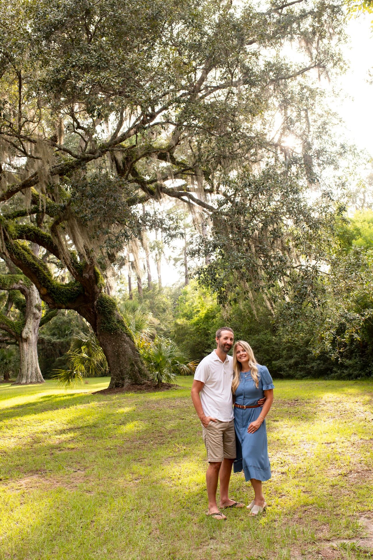 Couple embracing under a large moss-draped oak tree in a lush green park, man in white shirt and woman in blue dress.