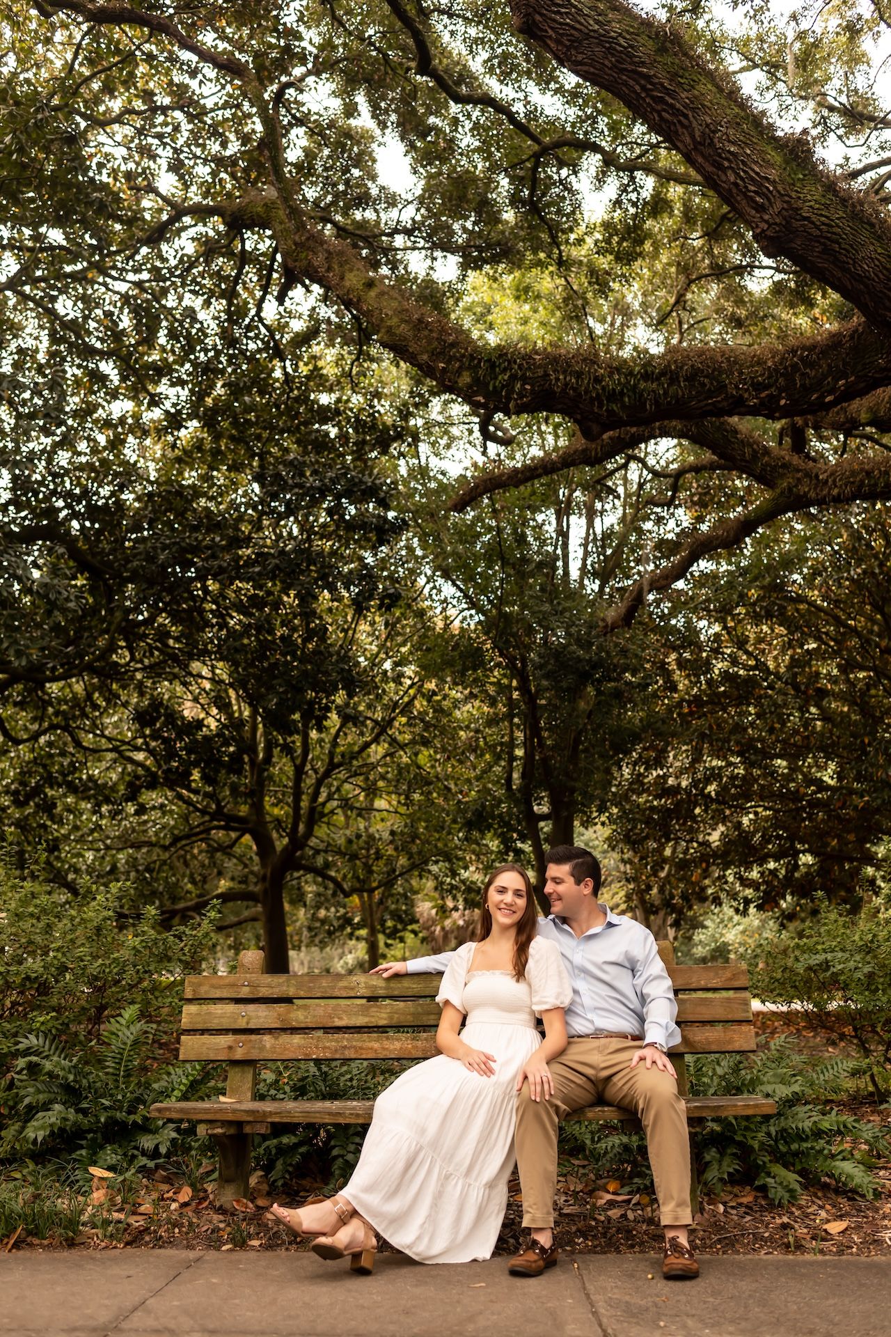 Couple sitting together on a wooden park bench surrounded by lush green trees, woman in white dress, man in light blue shirt.