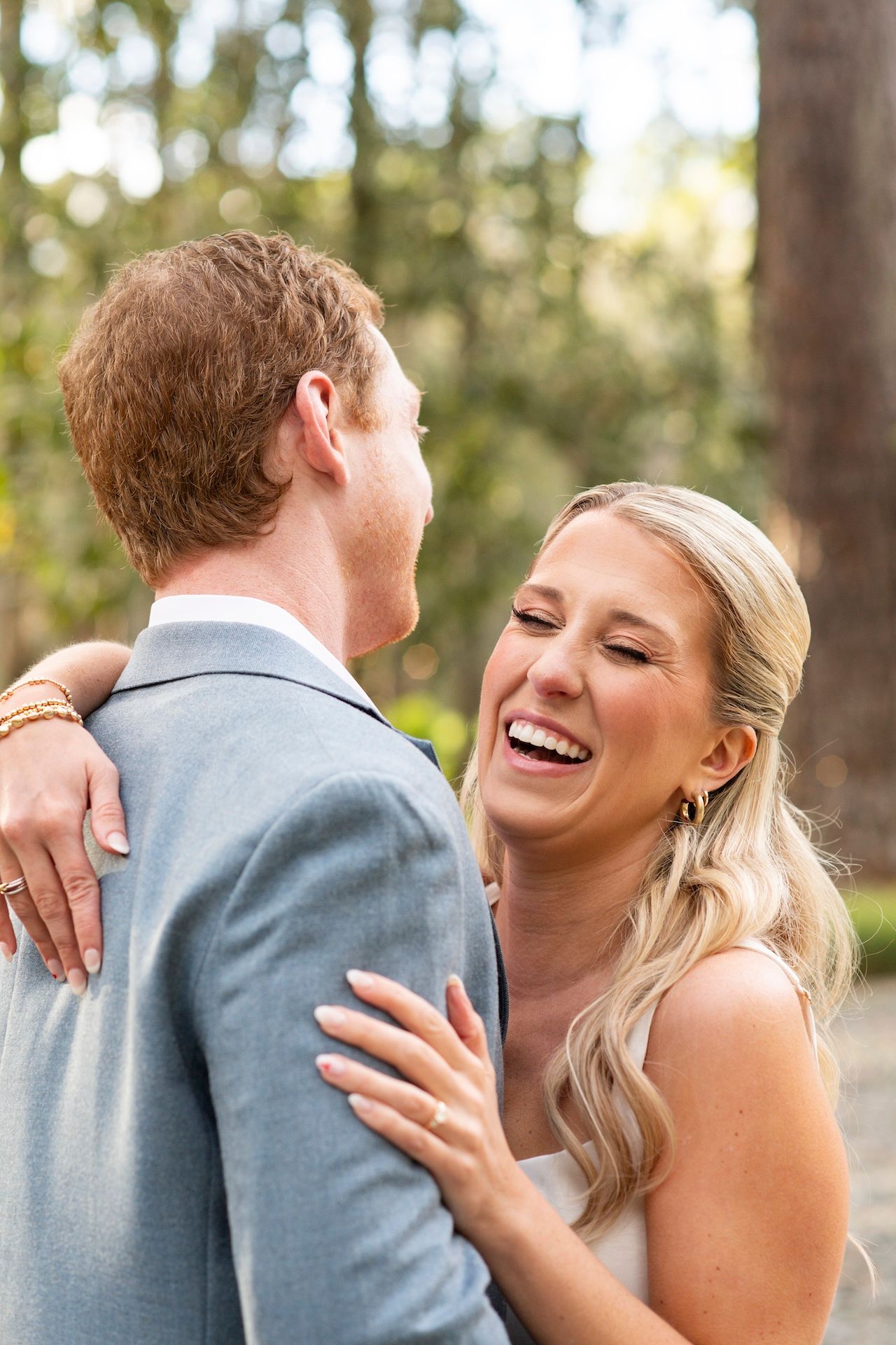A joyful bride laughs while embracing a groom in a light blue suit outdoors among trees on their wedding day.