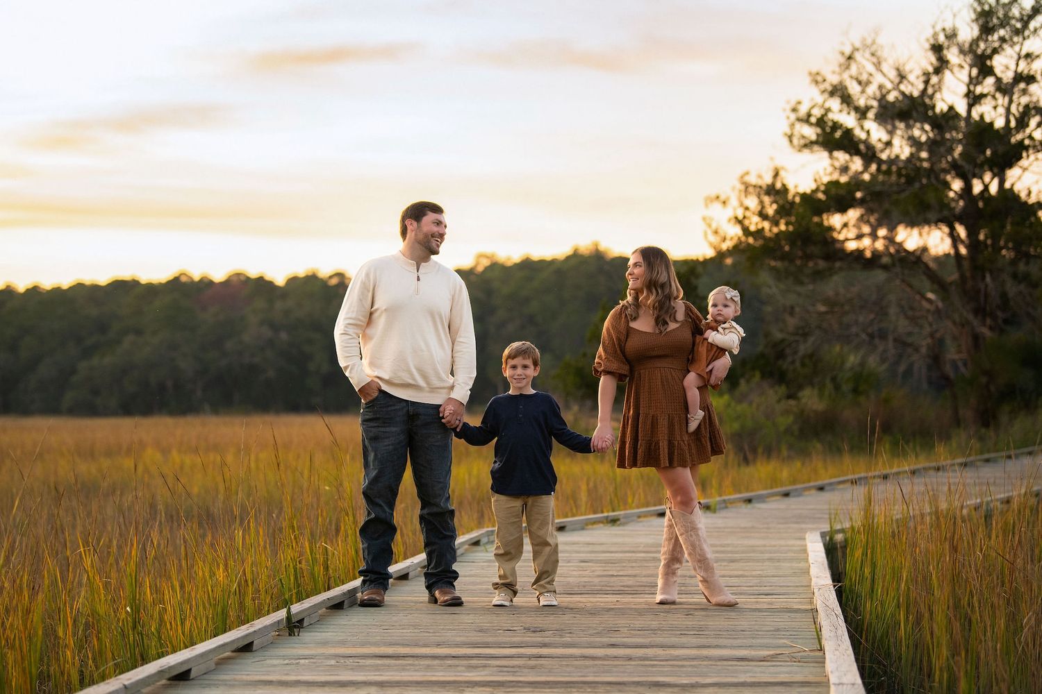 Family of four walks on a wooden boardwalk at sunset, surrounded by golden marsh grass and trees.