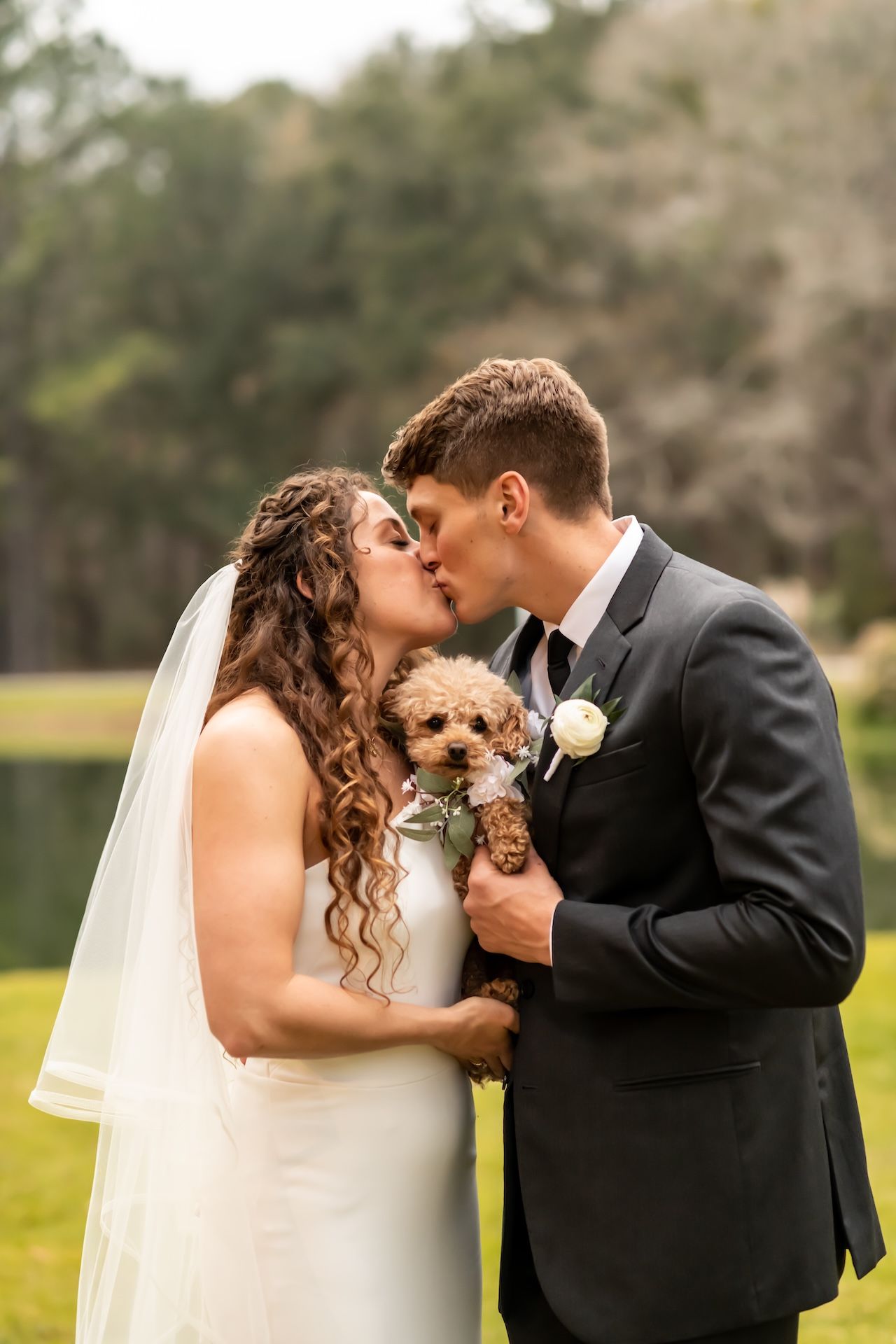 Bride and groom kissing while holding a small brown poodle wearing floral collar on their wedding day outdoors.