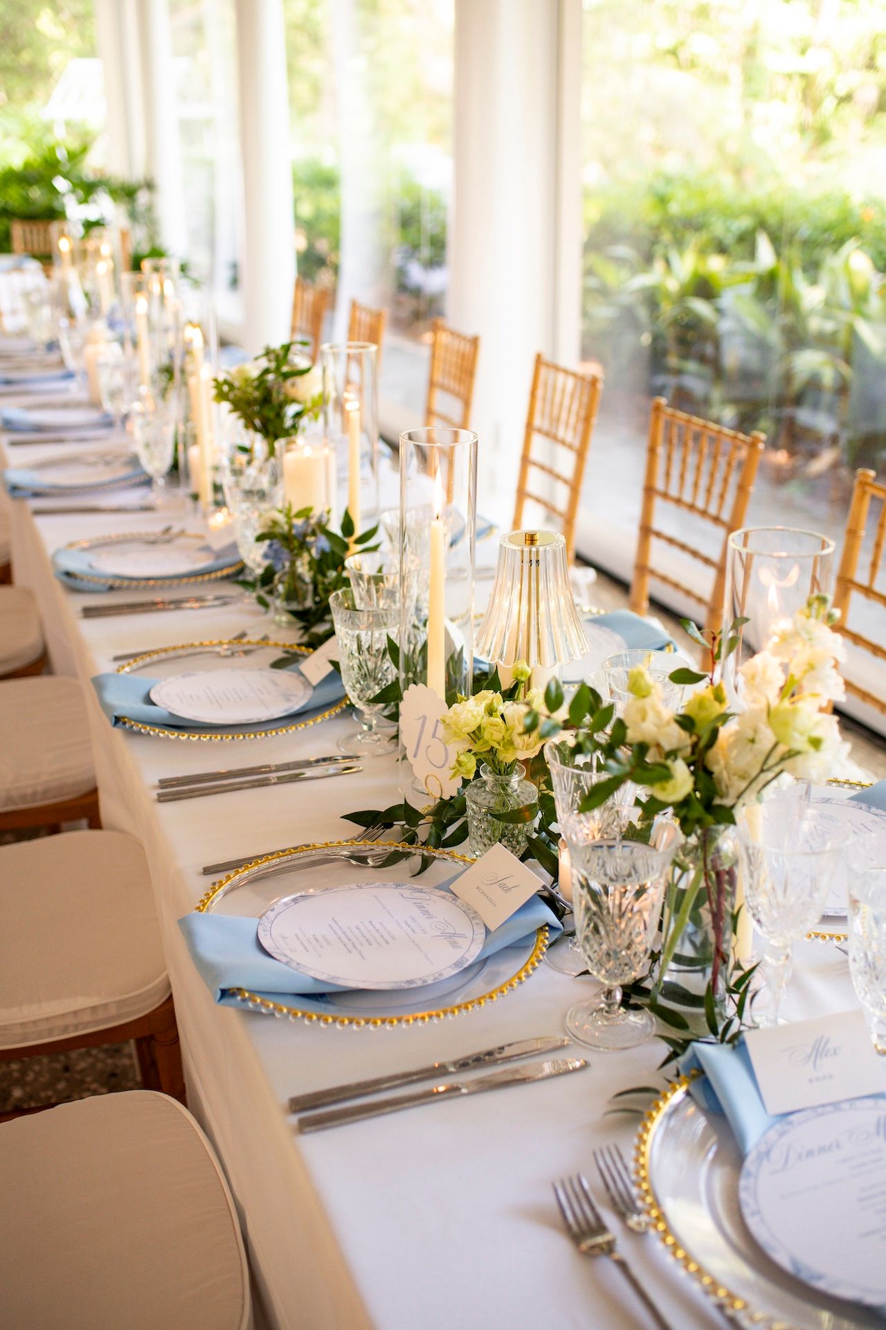 Elegant wedding reception table with blue napkins, gold chargers, white floral centerpieces, and candles on a white tablecloth.