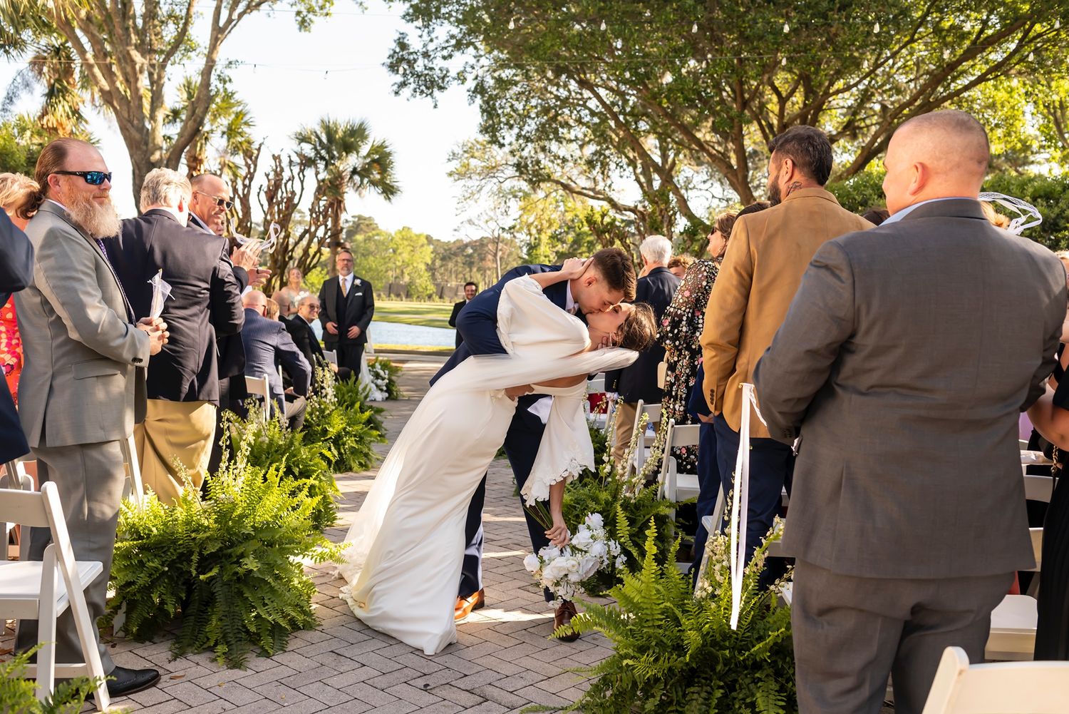 Wedding celebration under tree-lined path with people gathered around as a couple share a romantic dip during ceremony.