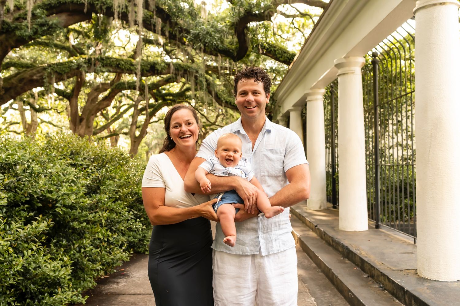 Happy people pose together on the steps of a white columned building with lush trees and landscaping in the background.