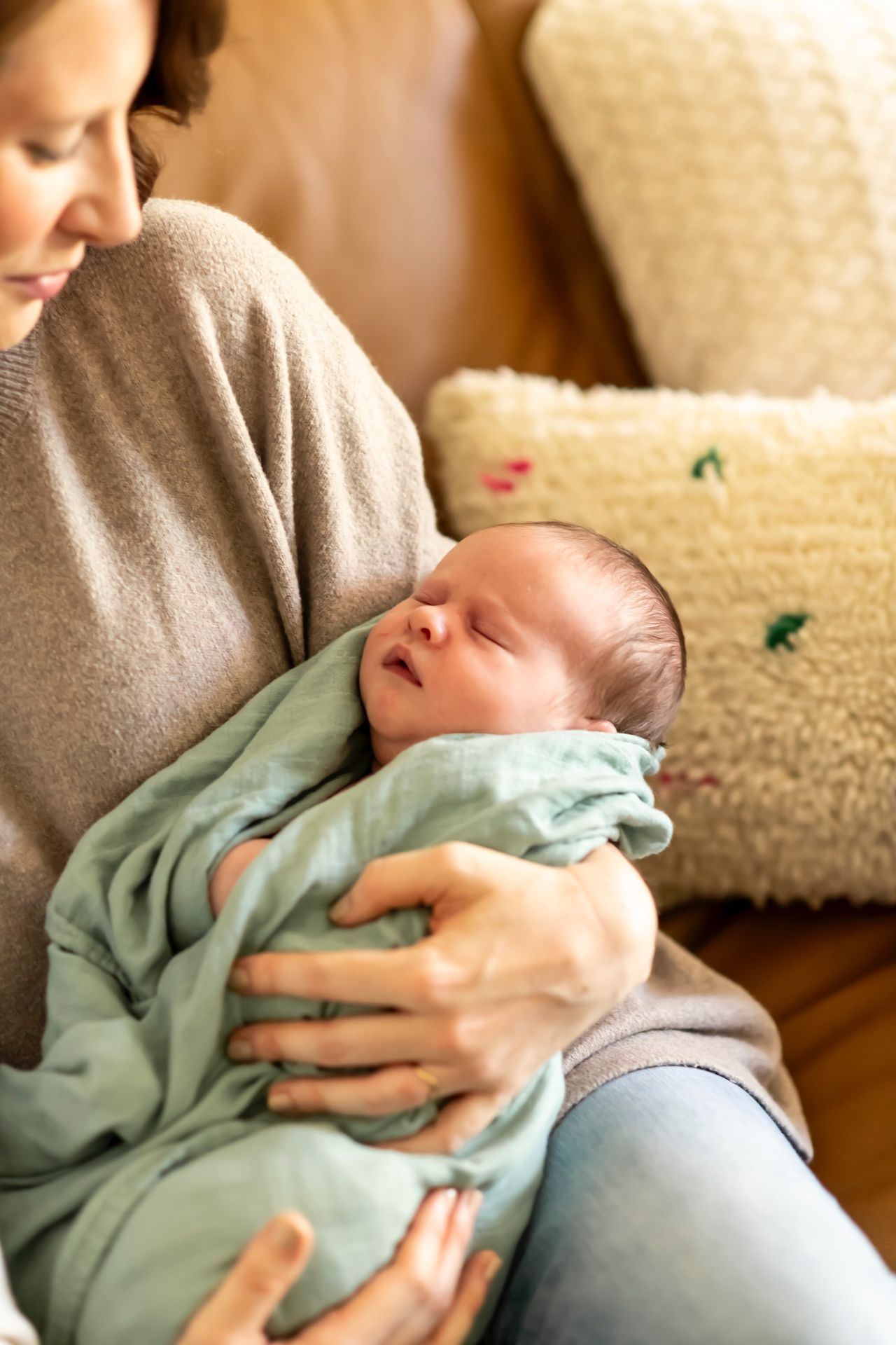 A newborn baby wrapped in a mint green blanket sleeps peacefully while being cradled in someone's arms on a beige sofa.
