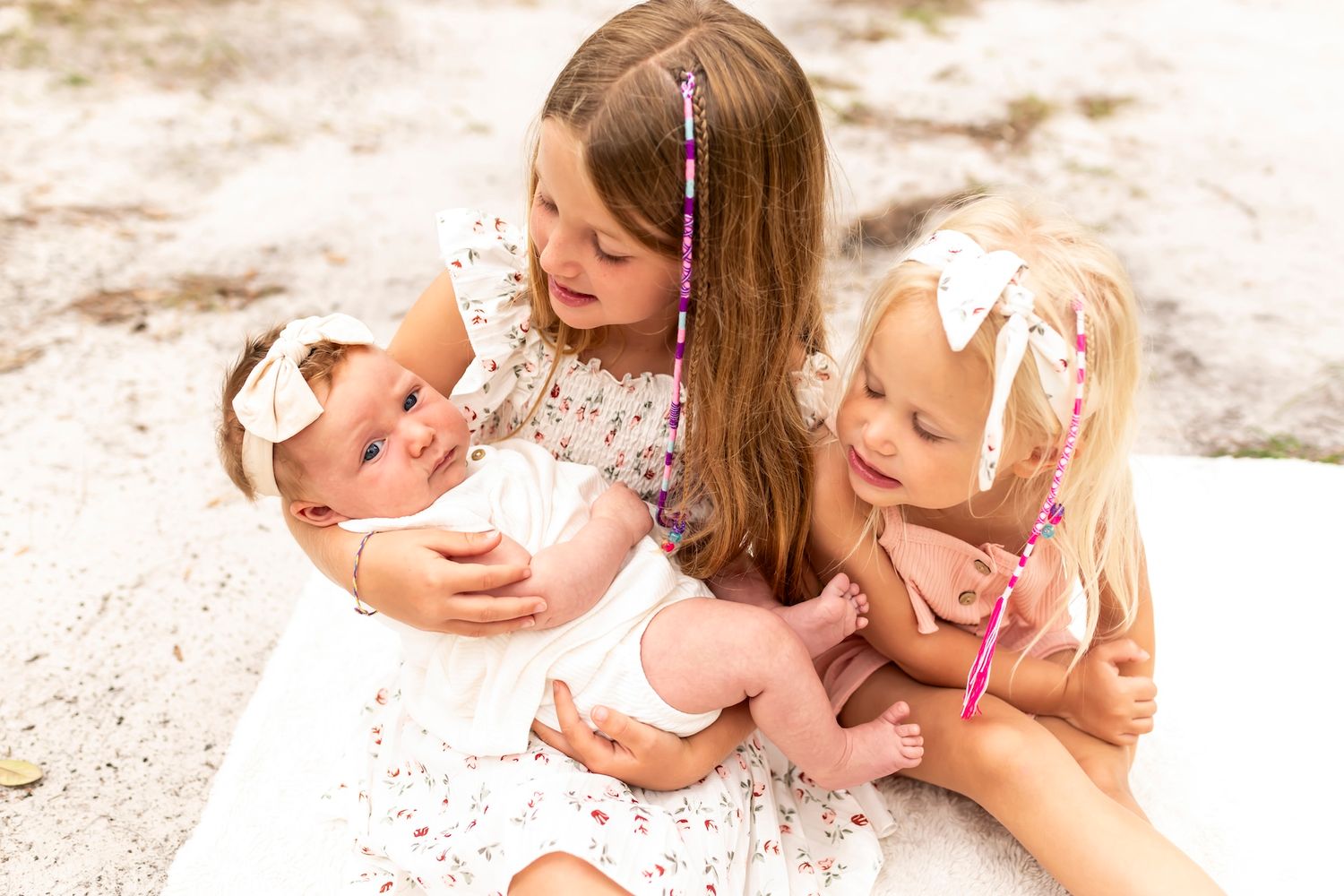 Three siblings in white lace dresses sitting together lovingly on a light sandy surface.