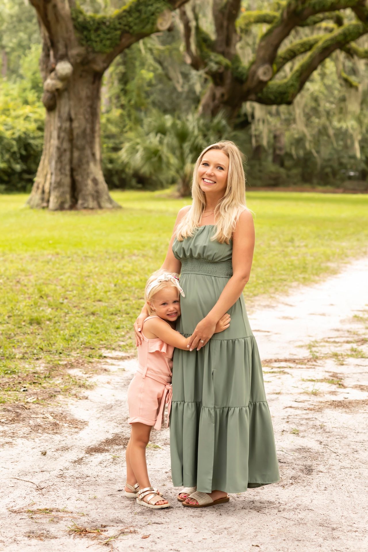 Family photoshoot in a park with moss-covered oak trees and a sandy path, wearing sage green and peach outfits.