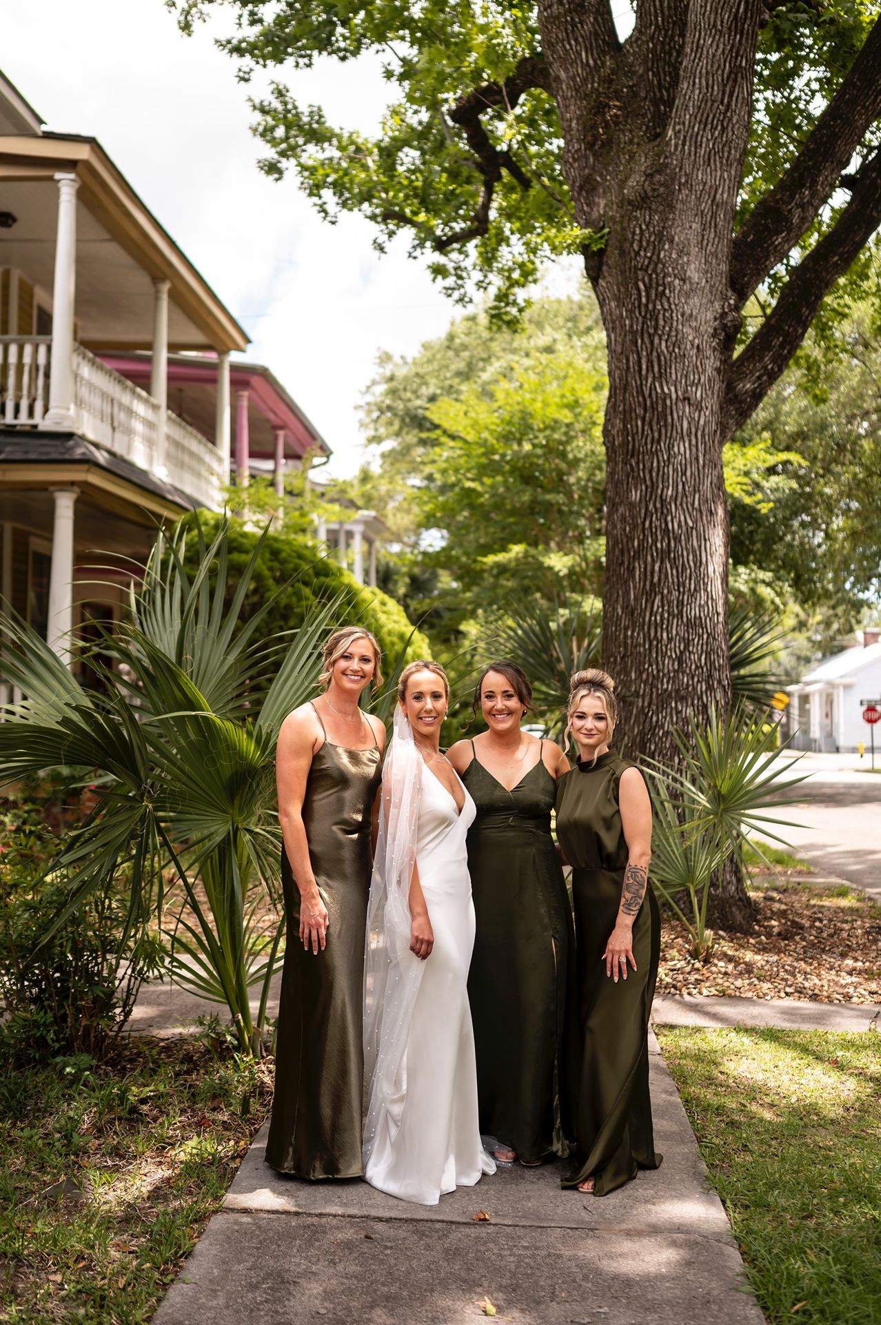Four women in formal dresses pose together on a garden path in front of a historic Southern-style house with palms.