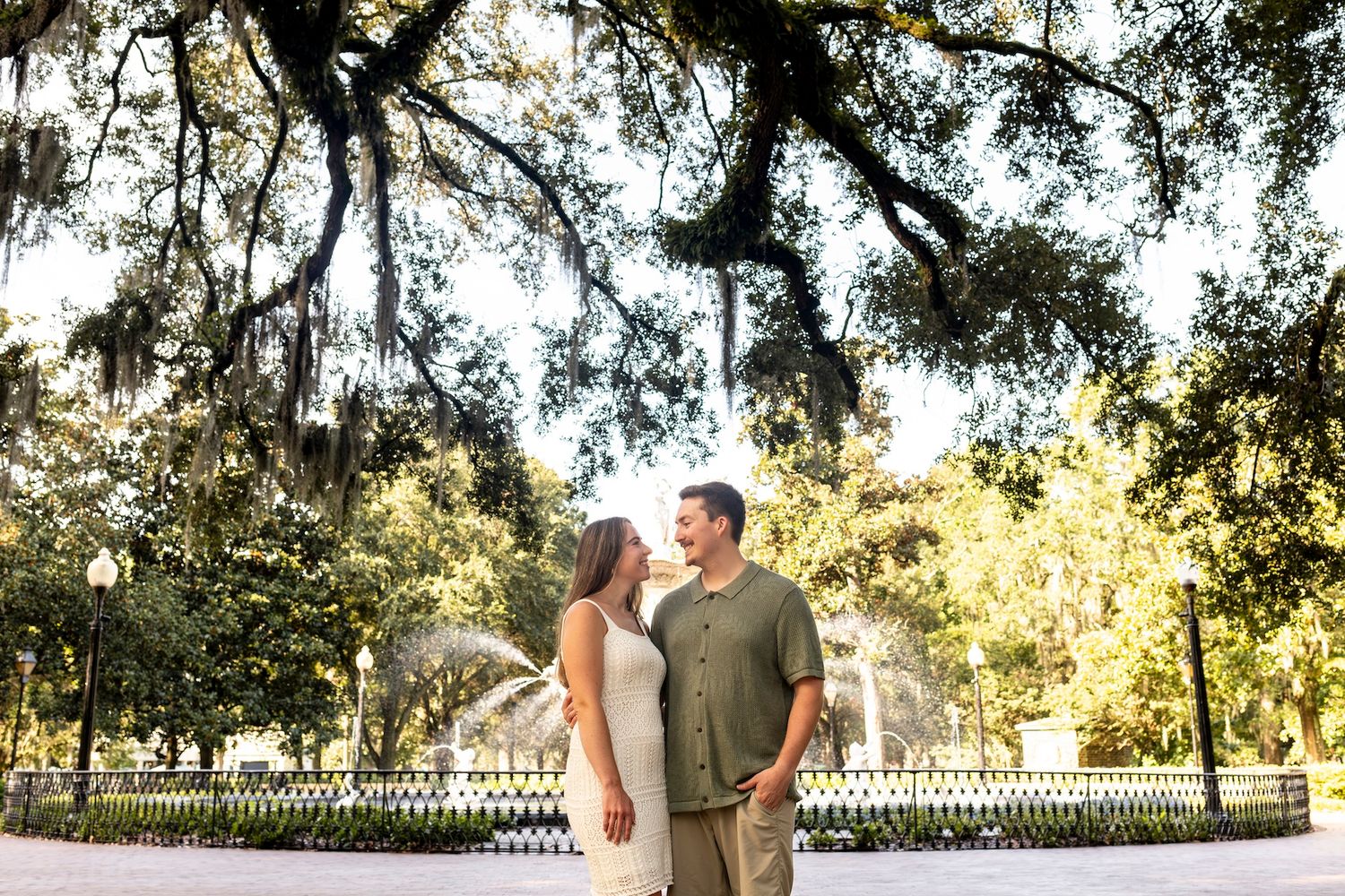 A couple in casual attire shares a romantic moment under Spanish moss draped trees in a Southern park setting.