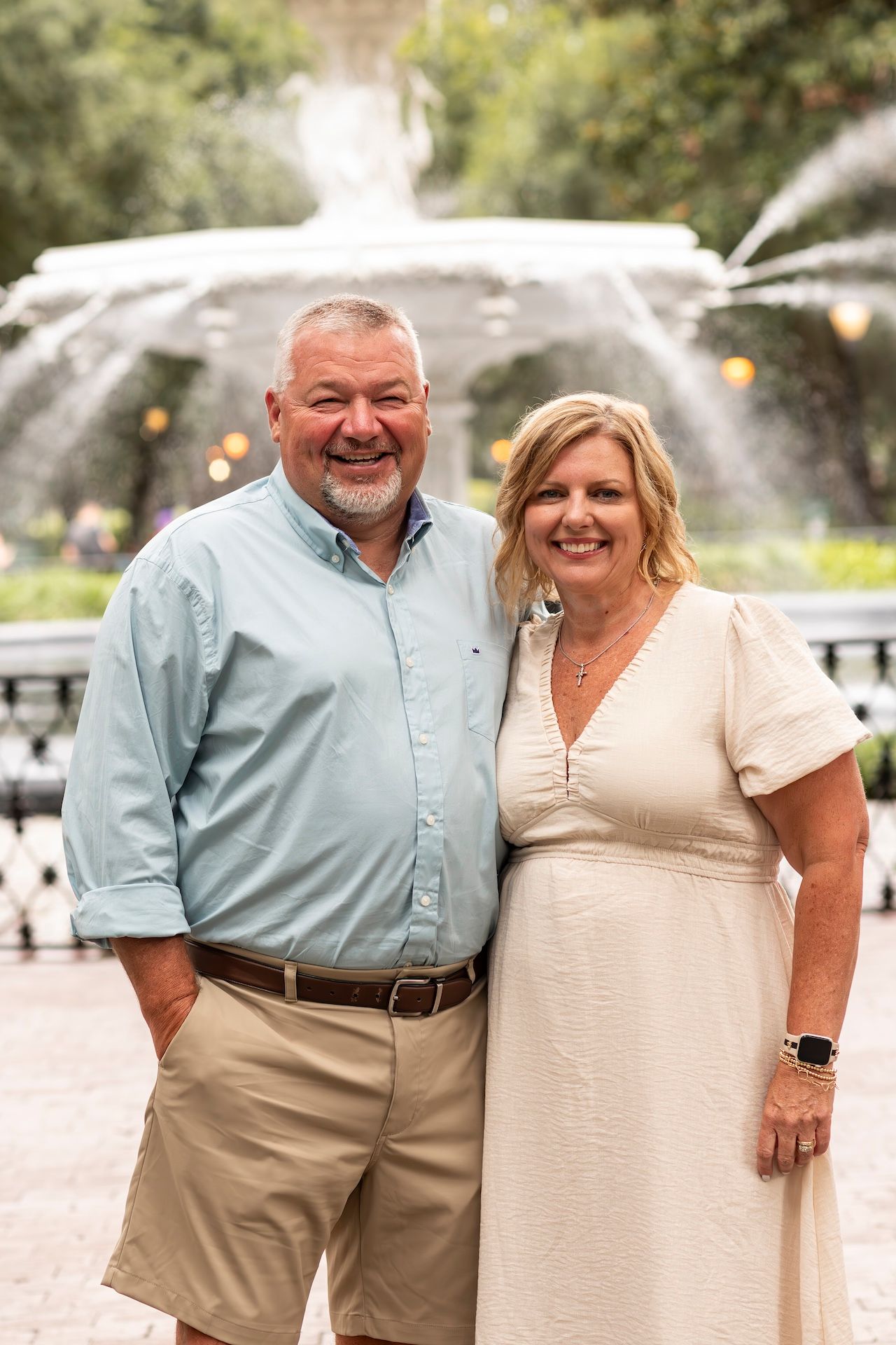 A couple poses in front of a fountain in a garden setting on a sunny day.