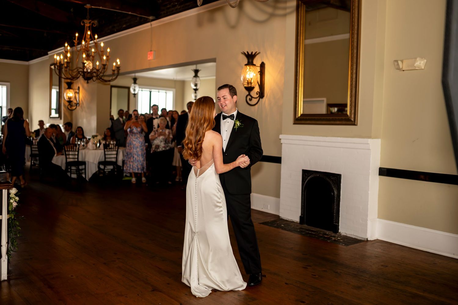 A couple shares their first dance in an elegant ballroom with hardwood floors, chandelier and white fireplace.
