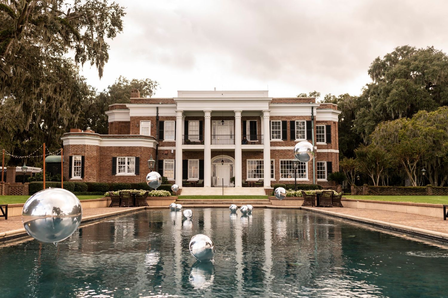 Elegant southern mansion with white columns and symmetrical design reflected in a pool with floating silver spheres.