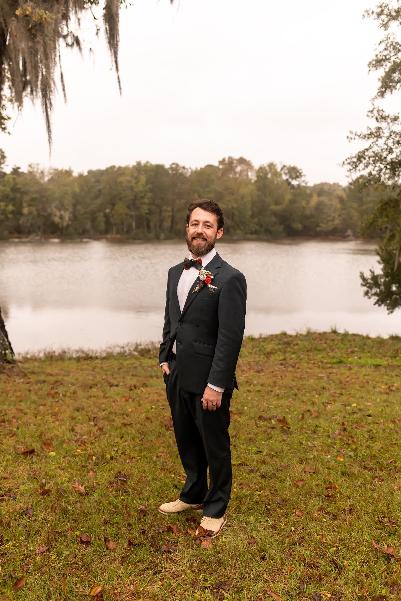 A groom in a black tuxedo with a beard stands on grassy lakeside with Spanish moss and trees in the background.