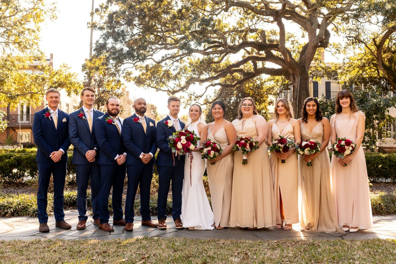 Wedding party wearing champagne and navy blue attire poses outdoors during sunset at elegant outdoor ceremony.