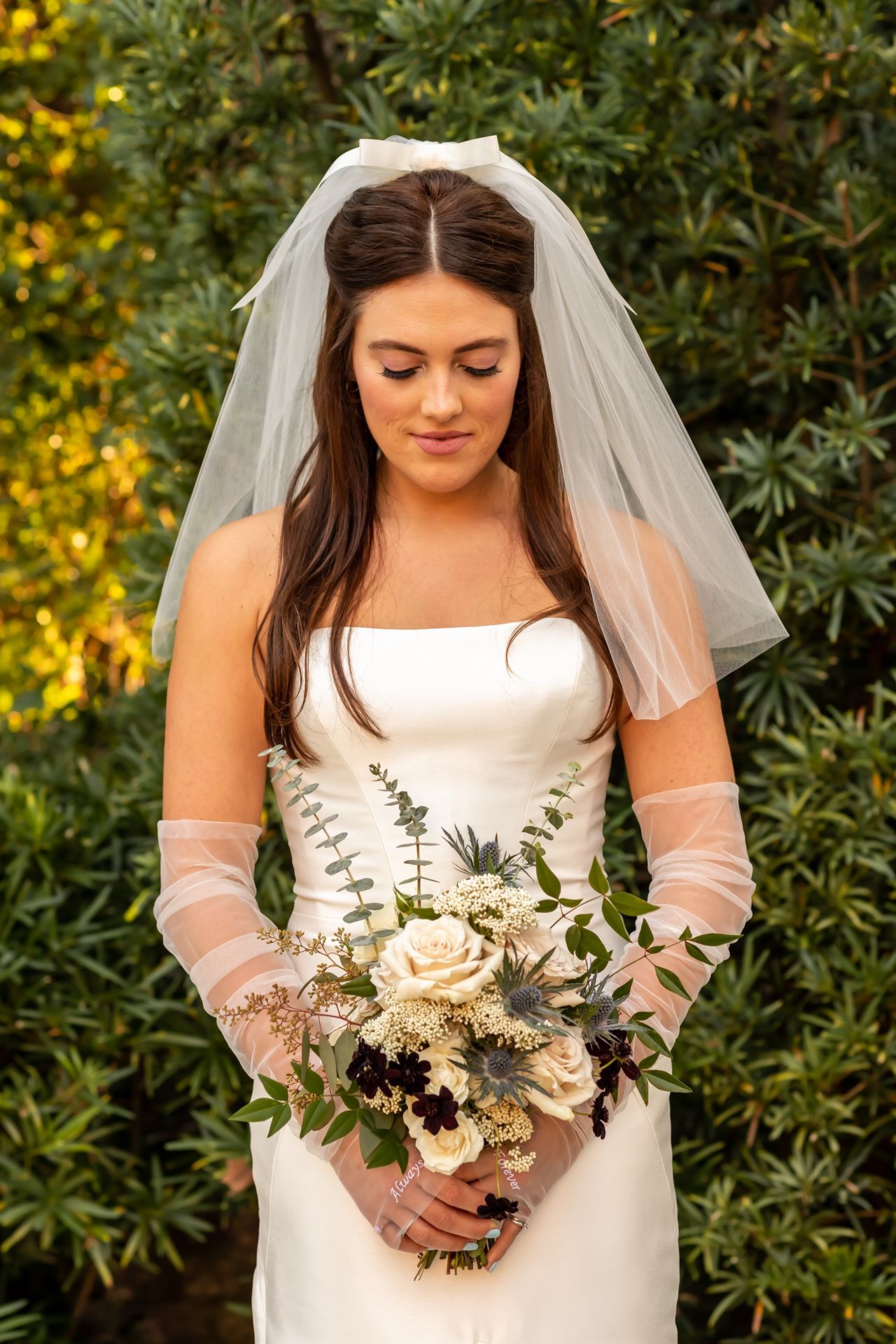 A bride in a white strapless dress and veil holds a rustic bouquet of cream roses and greenery against a garden backdrop.