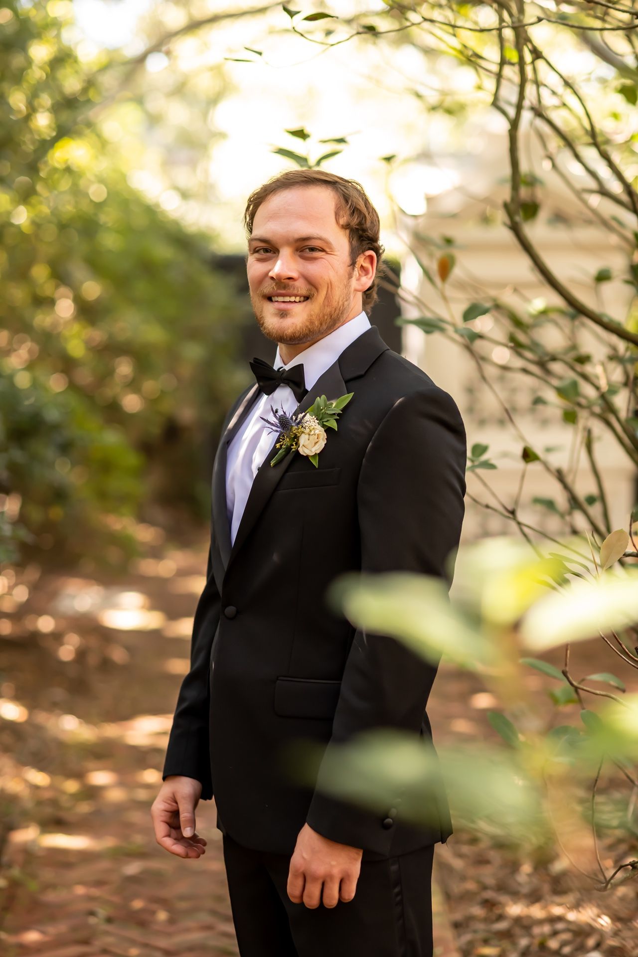 A person wearing a formal black tuxedo and boutonniere stands outdoors in a wooded setting with soft golden lighting.