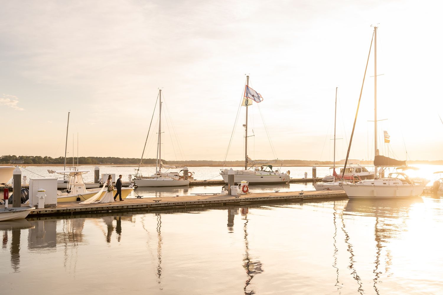 Sailboats docked at a marina during golden sunset with calm water reflections and American flag waving in the breeze.