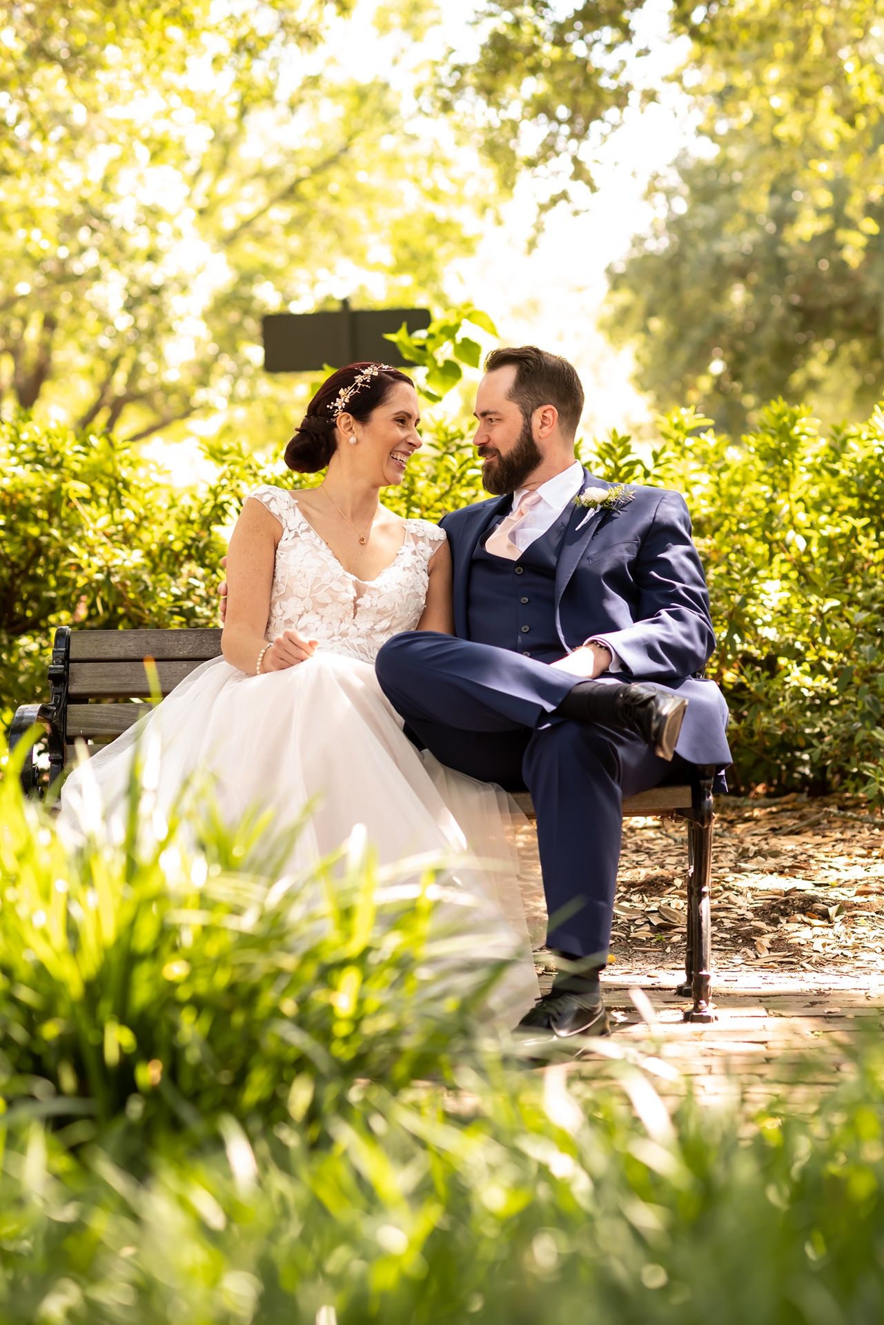 Newlyweds sit together on a park bench surrounded by lush greenery and dappled sunlight.