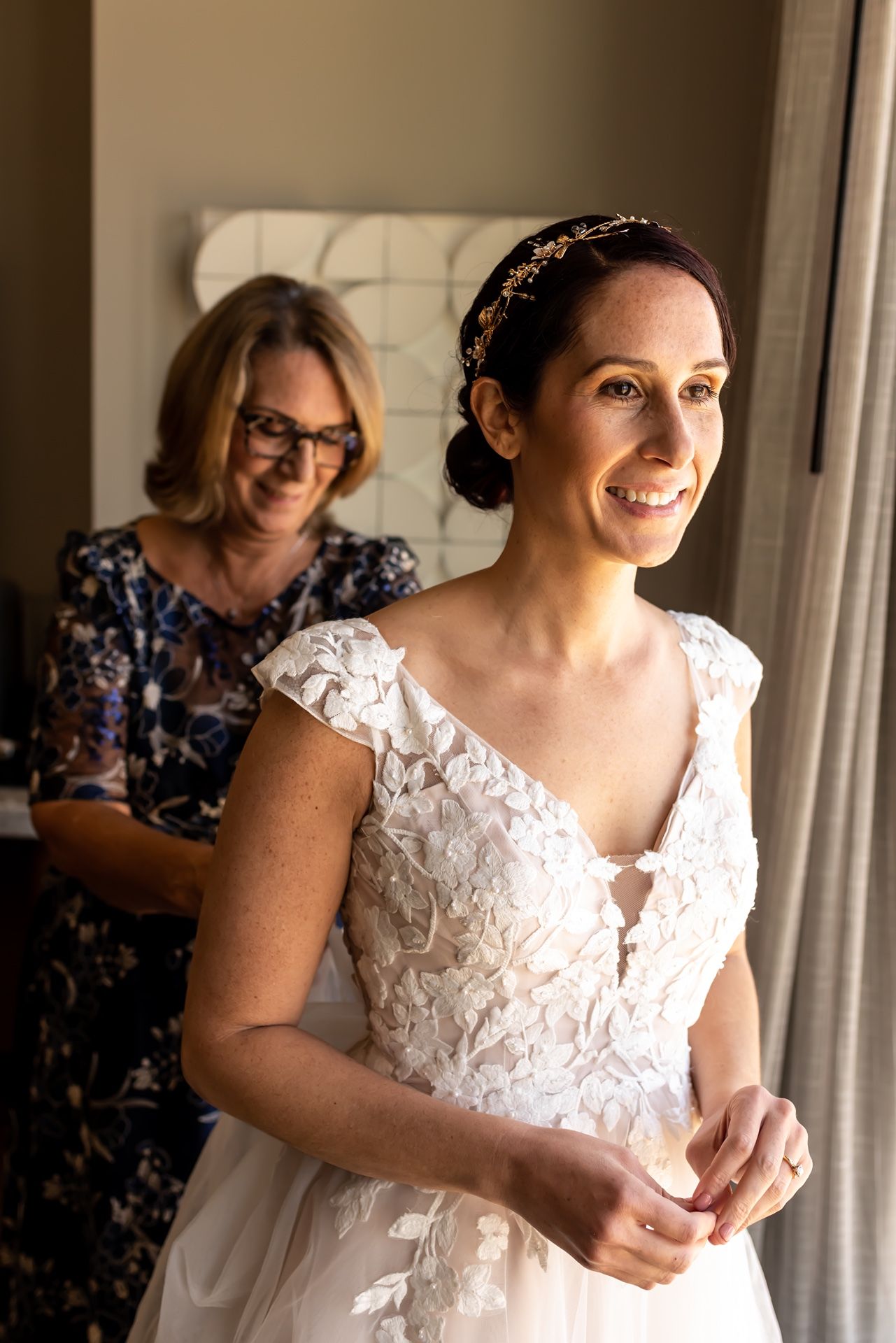 A bride in a white lace wedding dress smiles while getting ready with help in a softly lit room.
