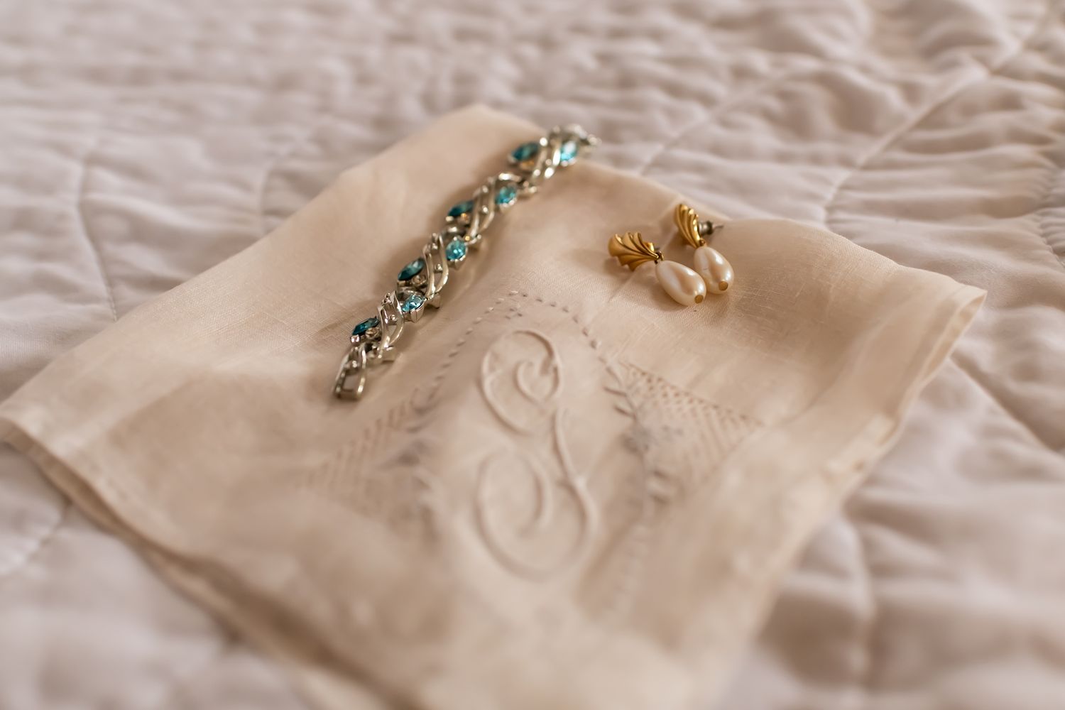 Vintage monogrammed handkerchief with delicate pearl jewelry and aqua bracelet laid on cream textured fabric.