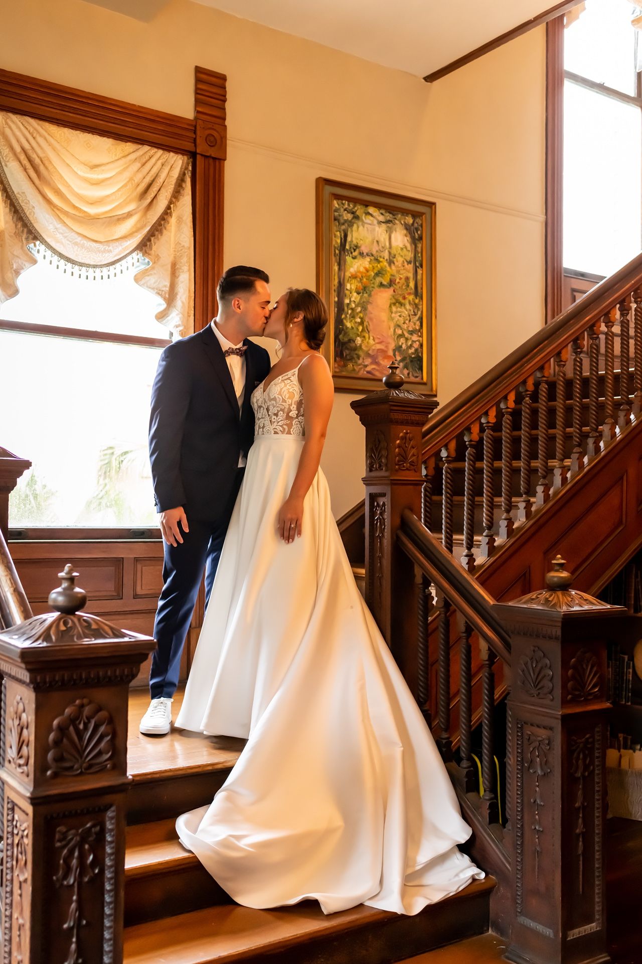 Elegant wedding couple standing on grand wooden staircase with ornate banister and flowing white bridal gown.