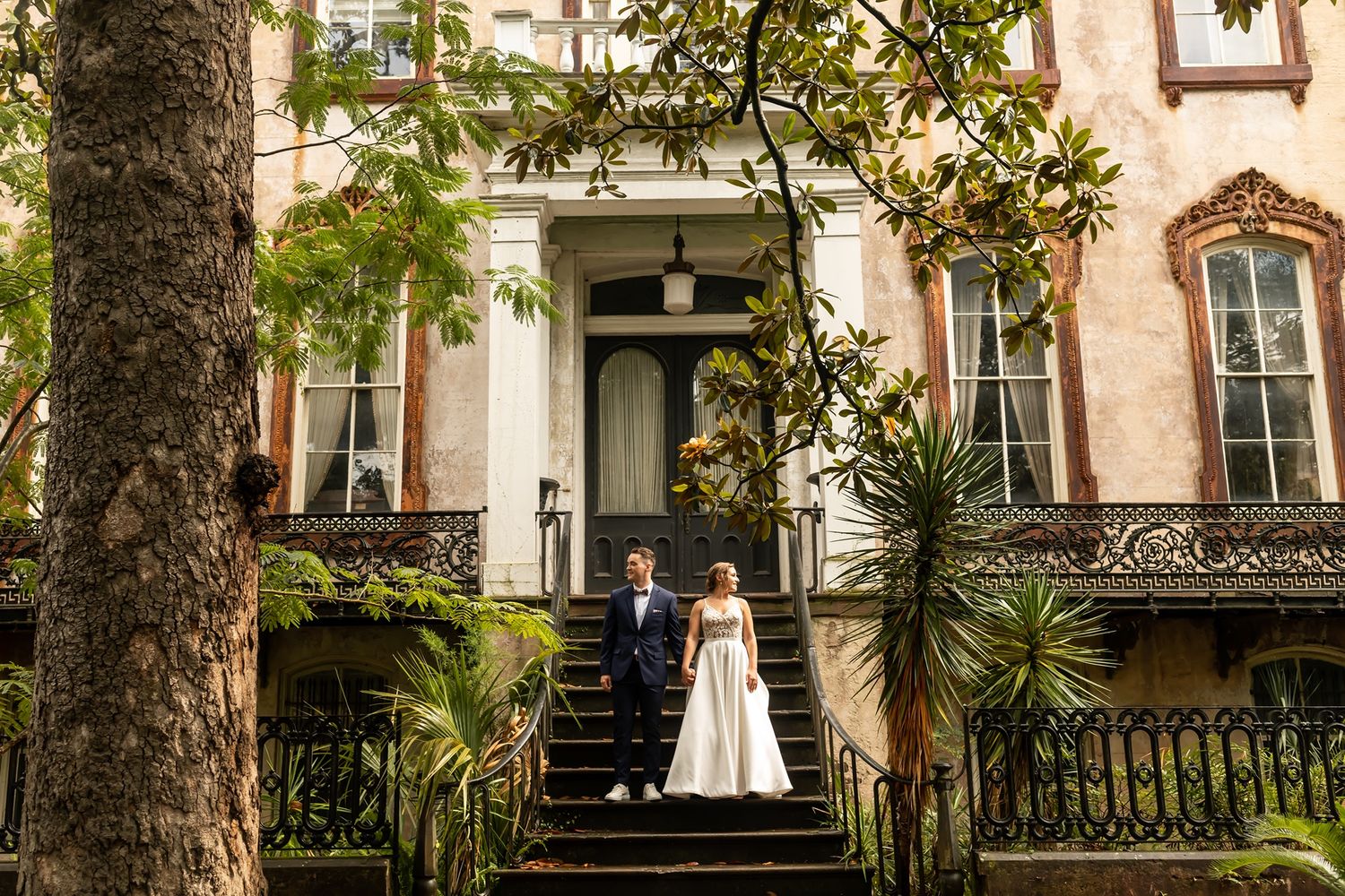 A couple stands on the ornate steps of a historic pink mansion in Savannah with palm trees and wrought iron details.
