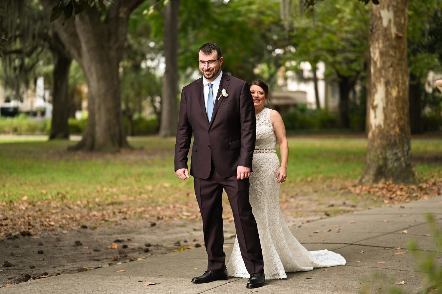 A couple in formal wedding attire poses together on a tree-lined path covered in fallen autumn leaves.