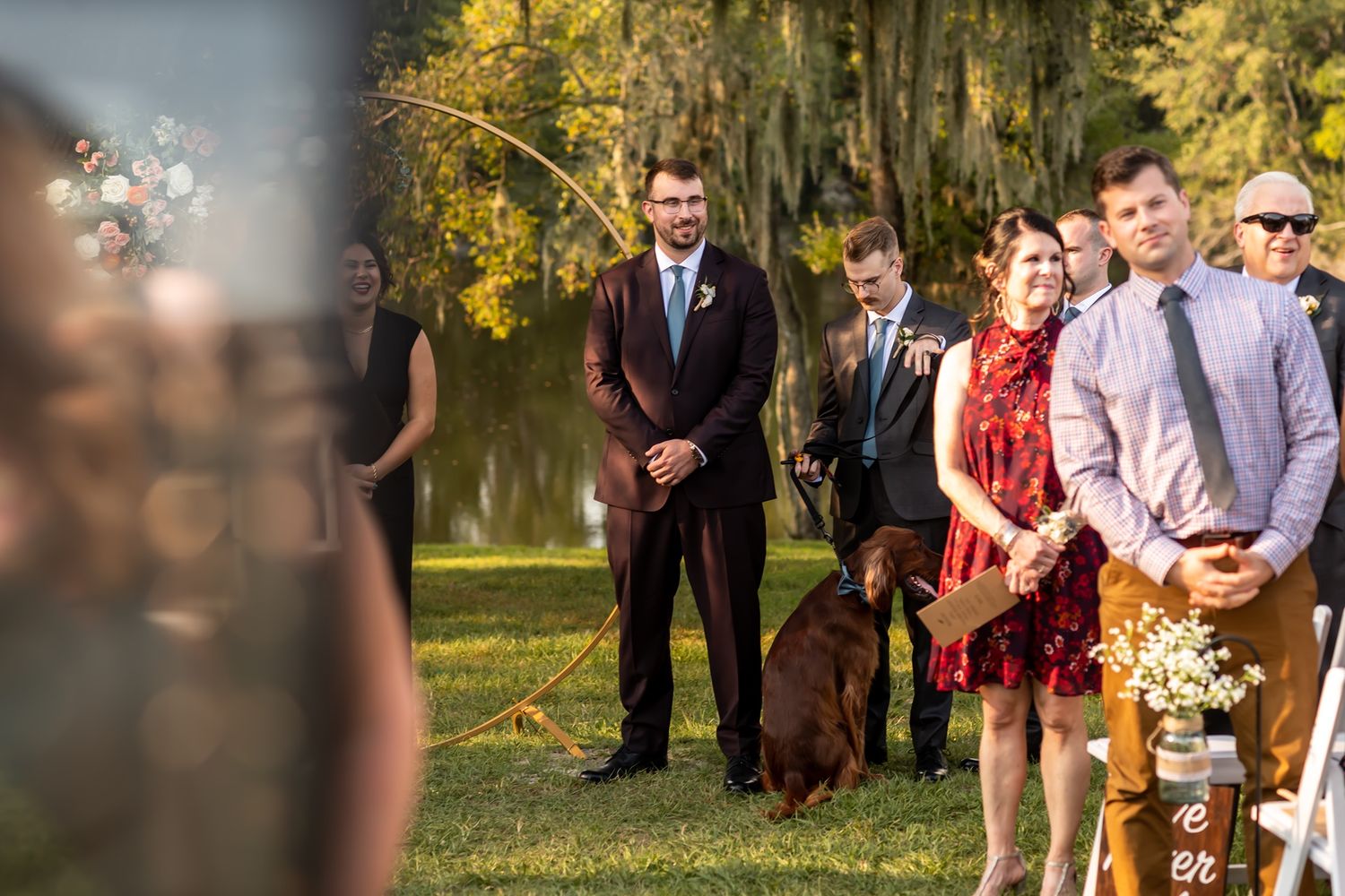 Wedding guests watch an outdoor ceremony under mossy oak trees in a scenic garden setting.
