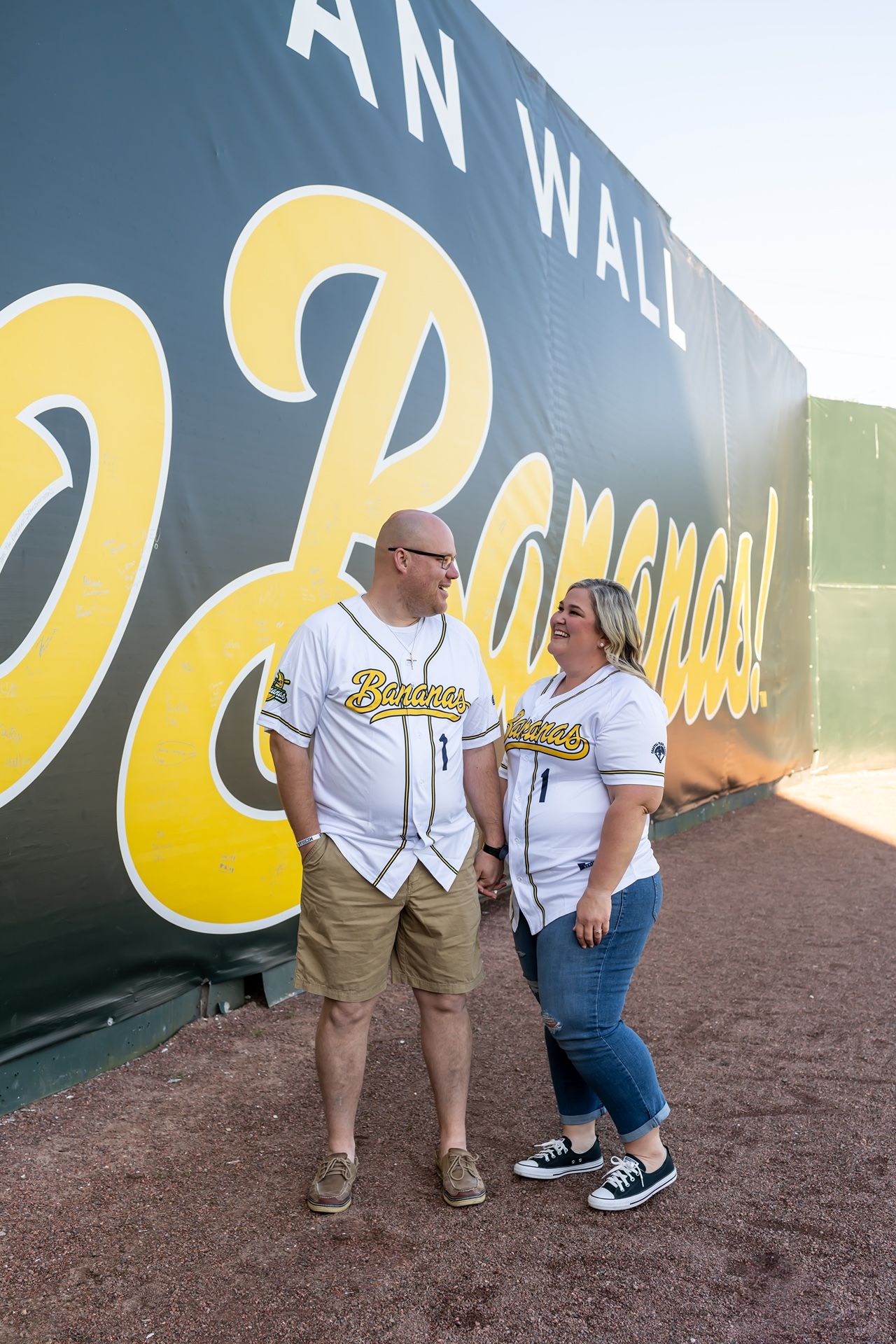 A couple in matching sports team apparel poses together in front of a large black wall with yellow lettering.
