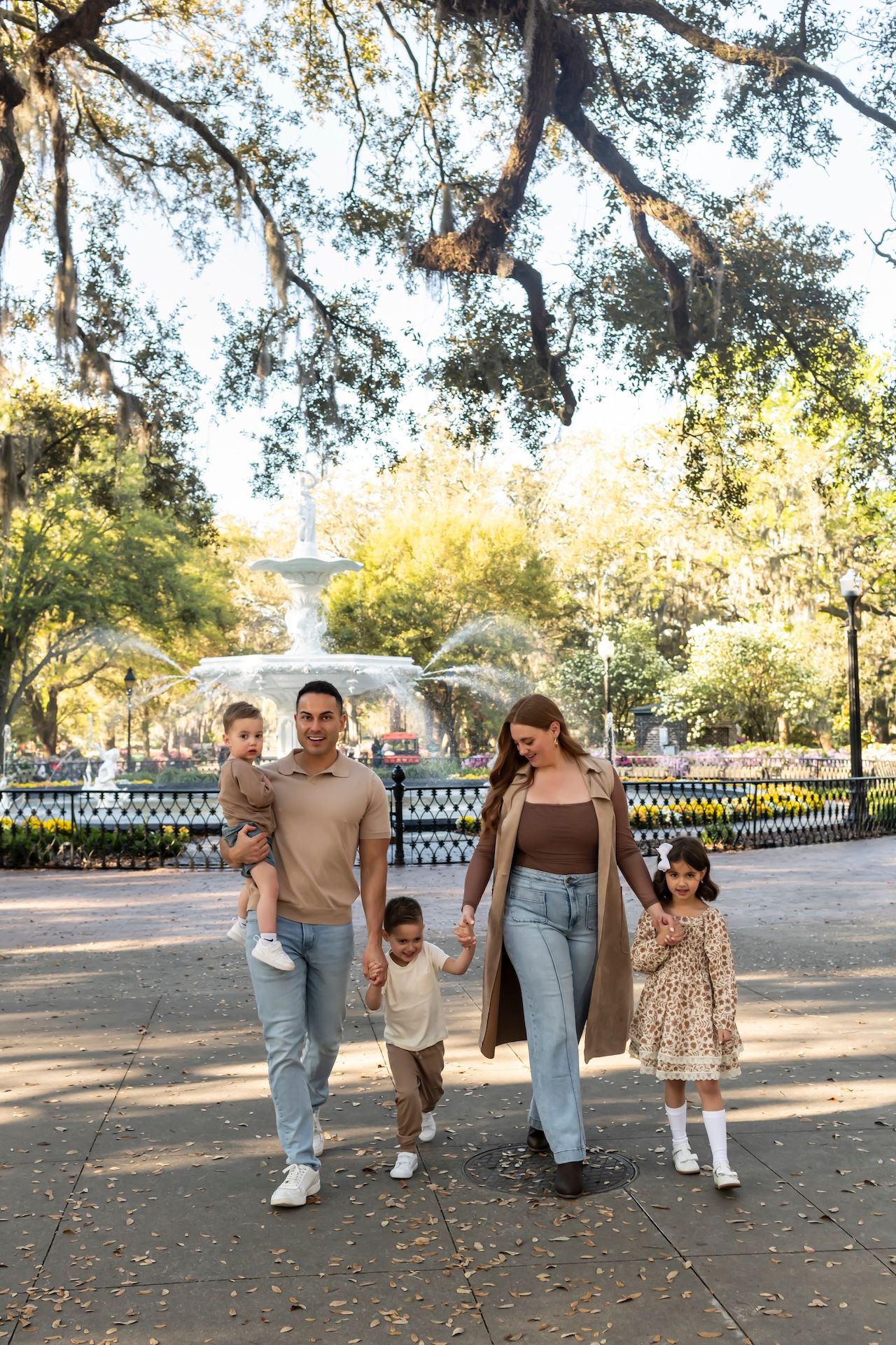 A family walks together on a tree-lined path in a park with a white fountain visible in the background.