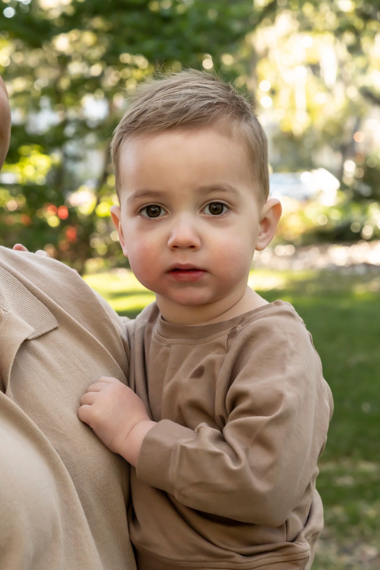 A young toddler wearing a beige sweater stands in a sunny outdoor park with bokeh effect in the background.