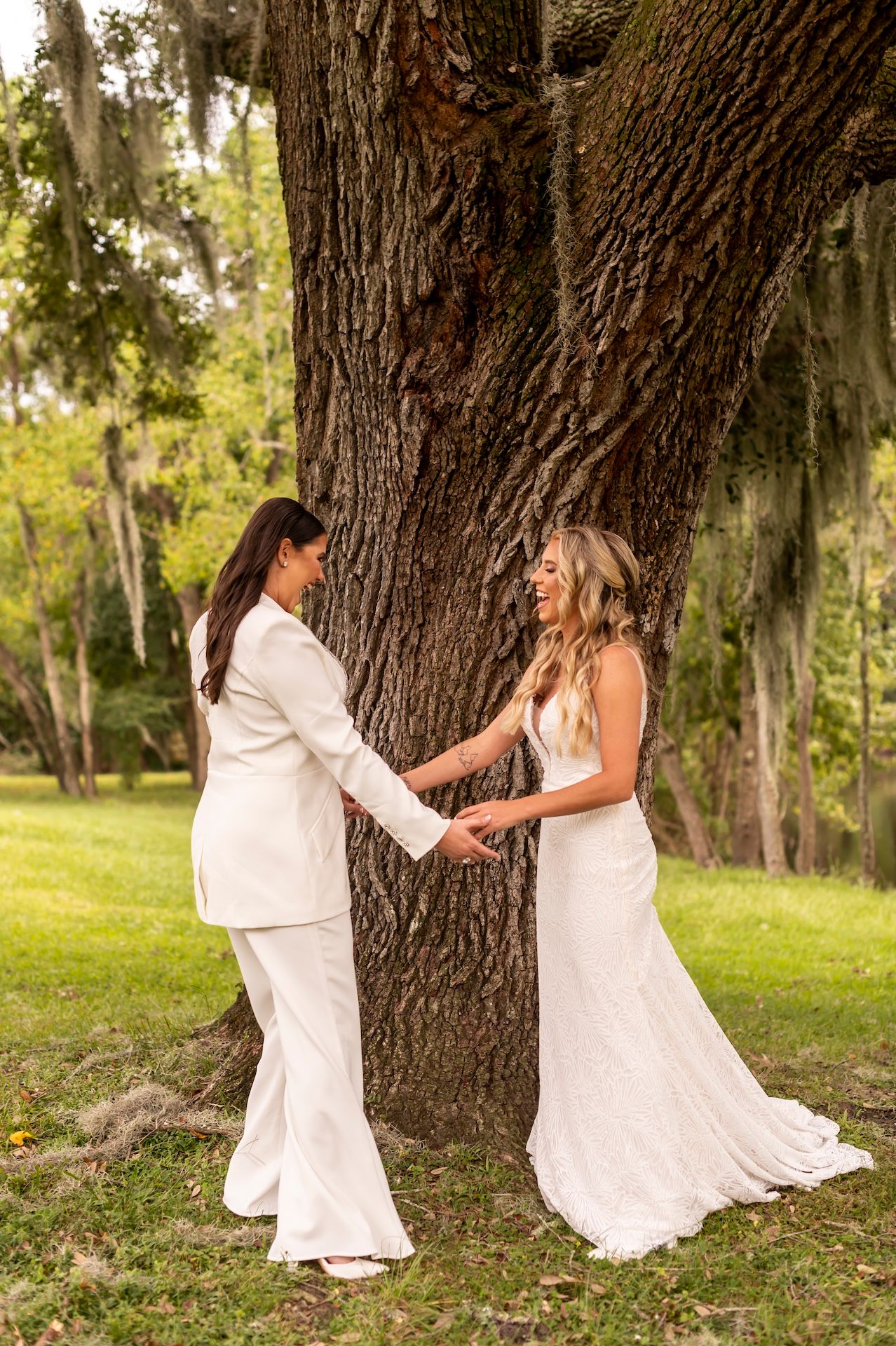 Two brides in elegant white gowns share an intimate moment beside a majestic tree during their outdoor wedding.