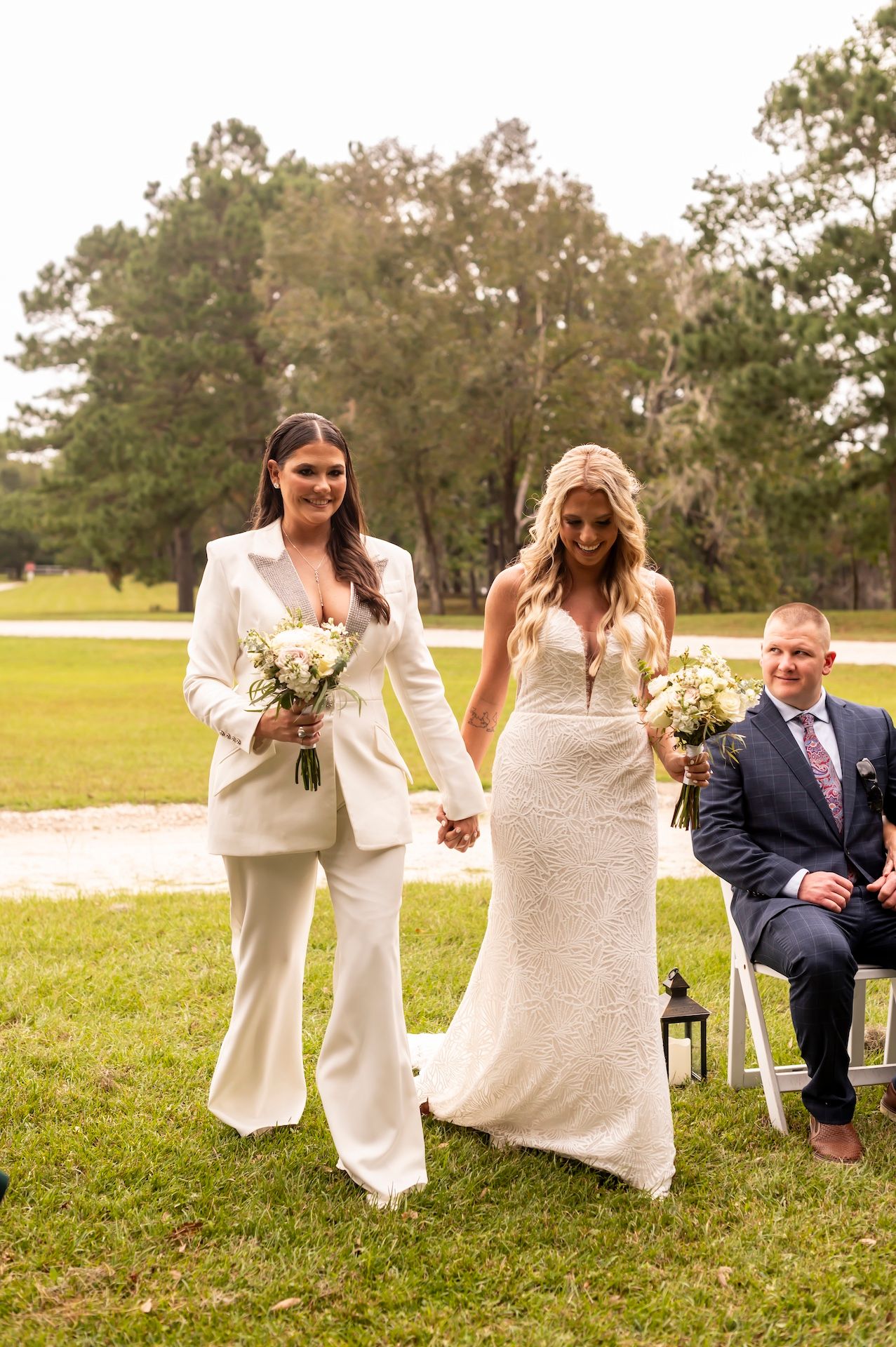 Two people in white wedding attire walk hand in hand on a grassy lawn while another person sits nearby.