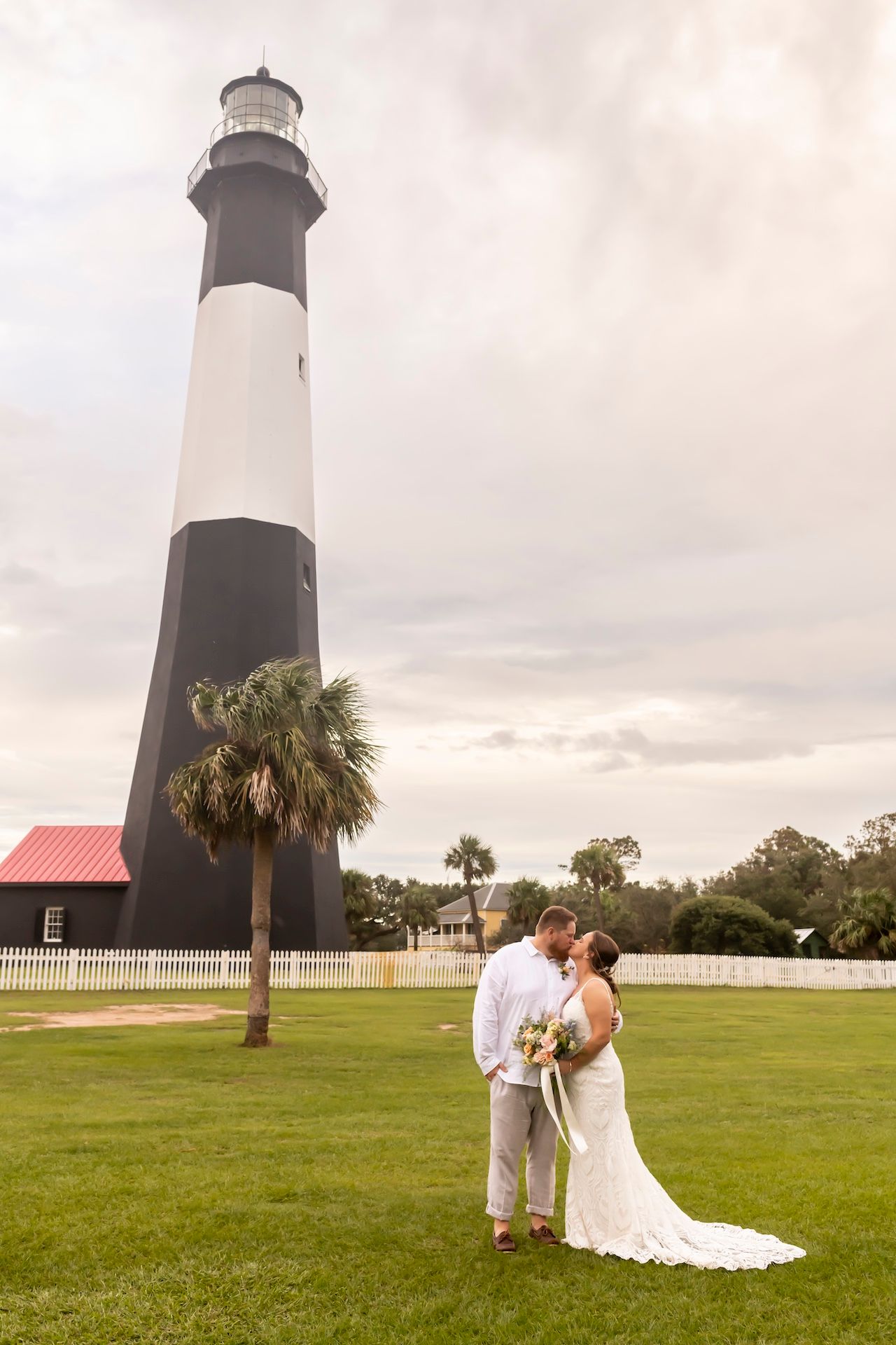 A tall black and white striped lighthouse stands behind a scenic grassy lawn with a palm tree against a cloudy sky.