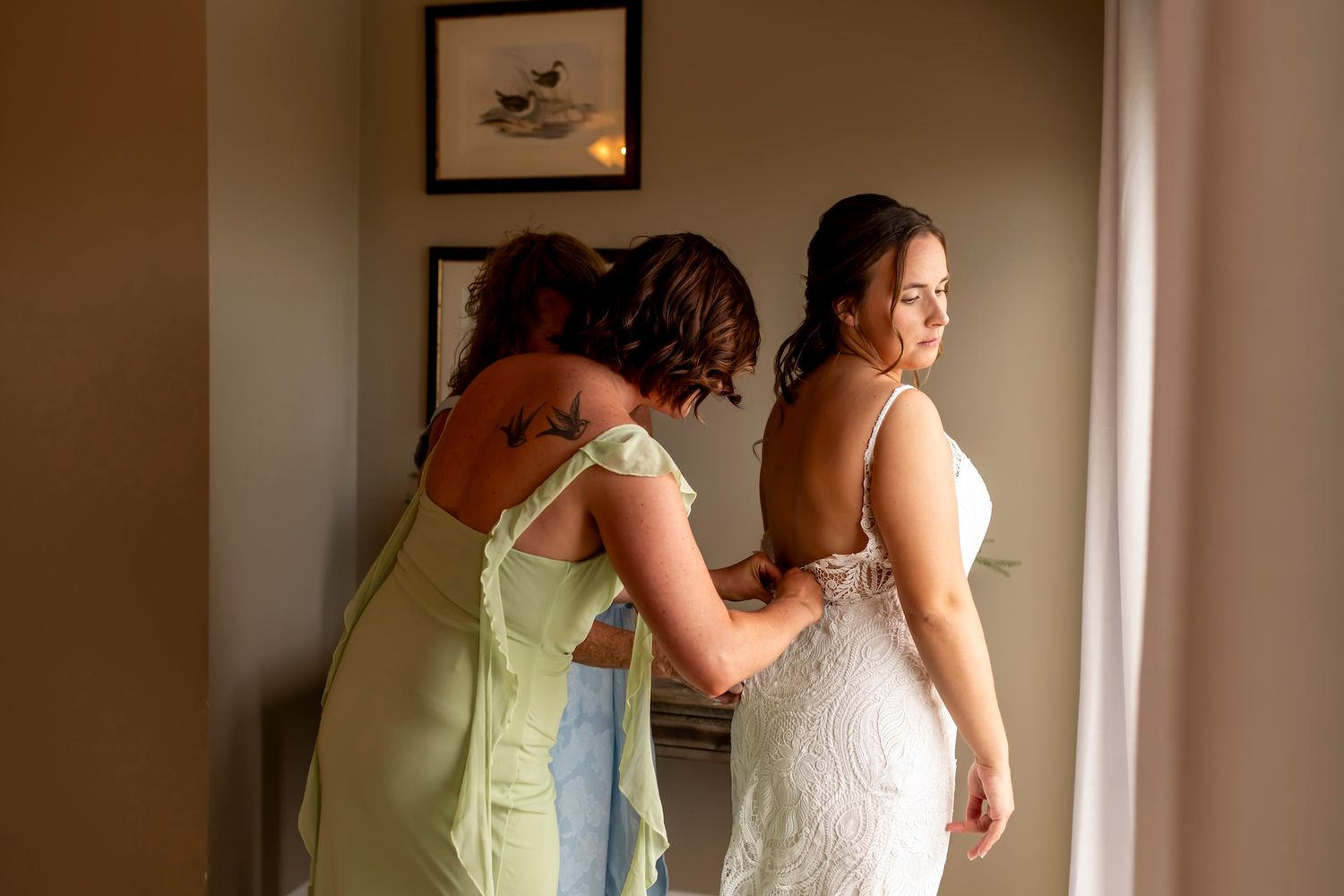 A bride in a white wedding dress gets her veil adjusted while standing near a window in soft natural light.
