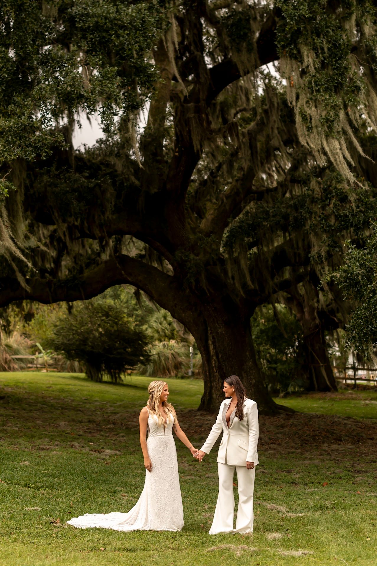 Two people in white outfits hold hands beneath a majestic Spanish moss-draped oak tree in a serene outdoor setting.