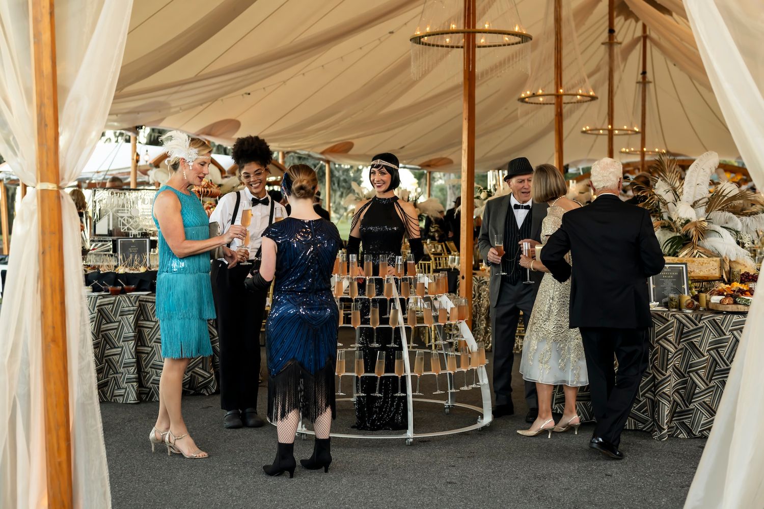 Event attendees gather under an elegant white tent around a tiered display of champagne glasses at an elegant party.