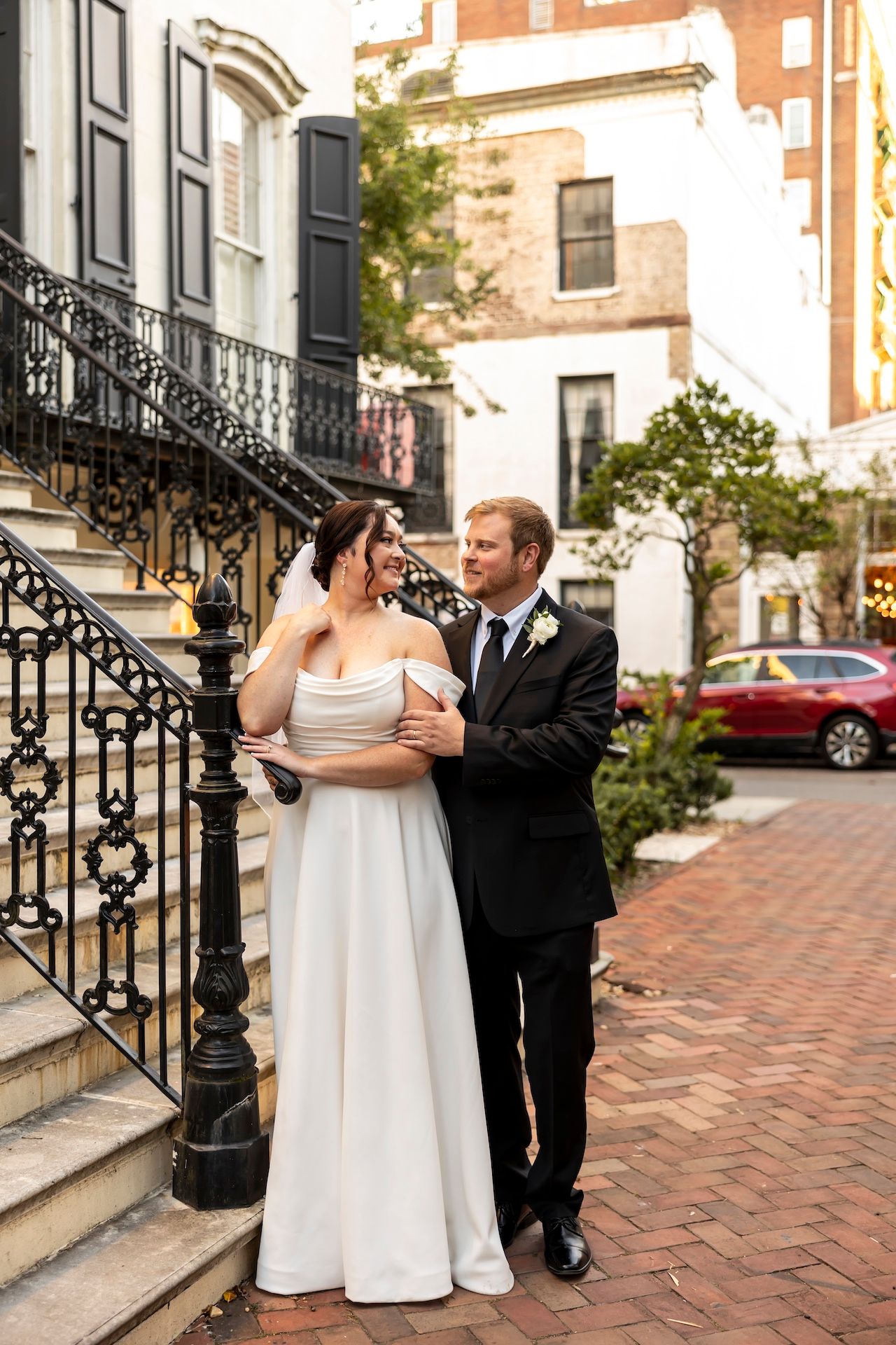 A couple stands on brick steps in front of historic Boston brownstones, with ornate black iron railings framing the scene.