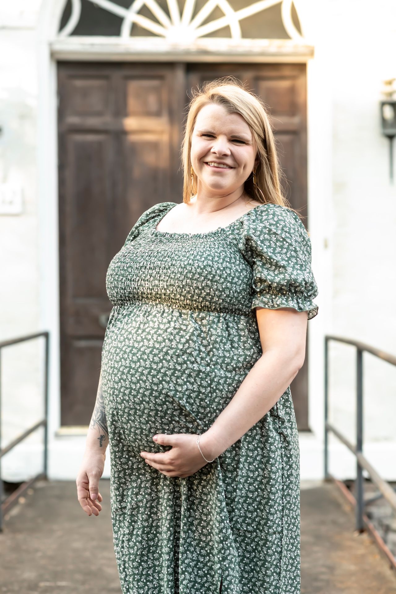 Someone in a green floral maternity dress stands outside a brown wooden door smiling and holding their pregnant belly.