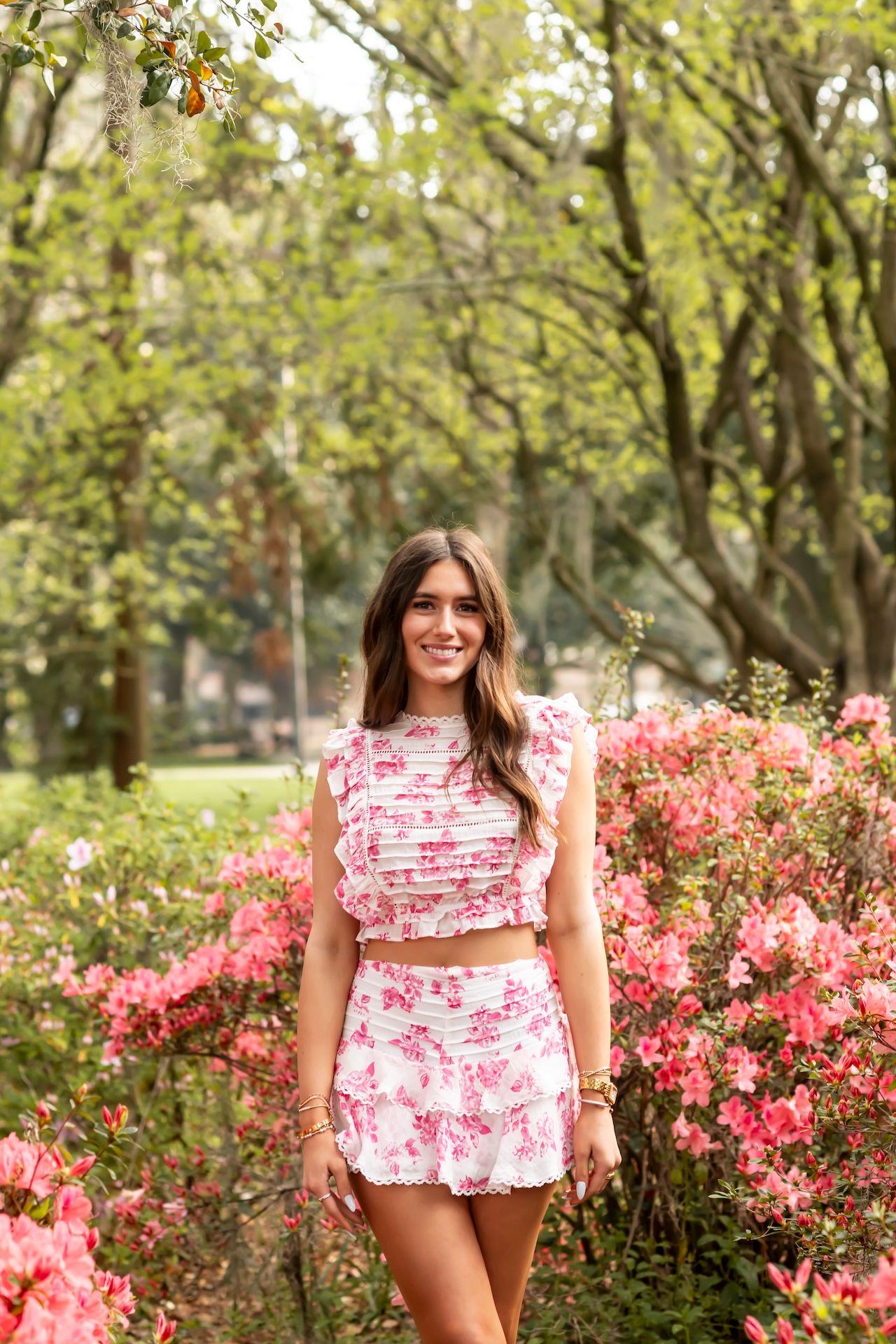 A pink floral two-piece outfit styled with a crop top and skirt stands out against vibrant azalea bushes in a garden setting.