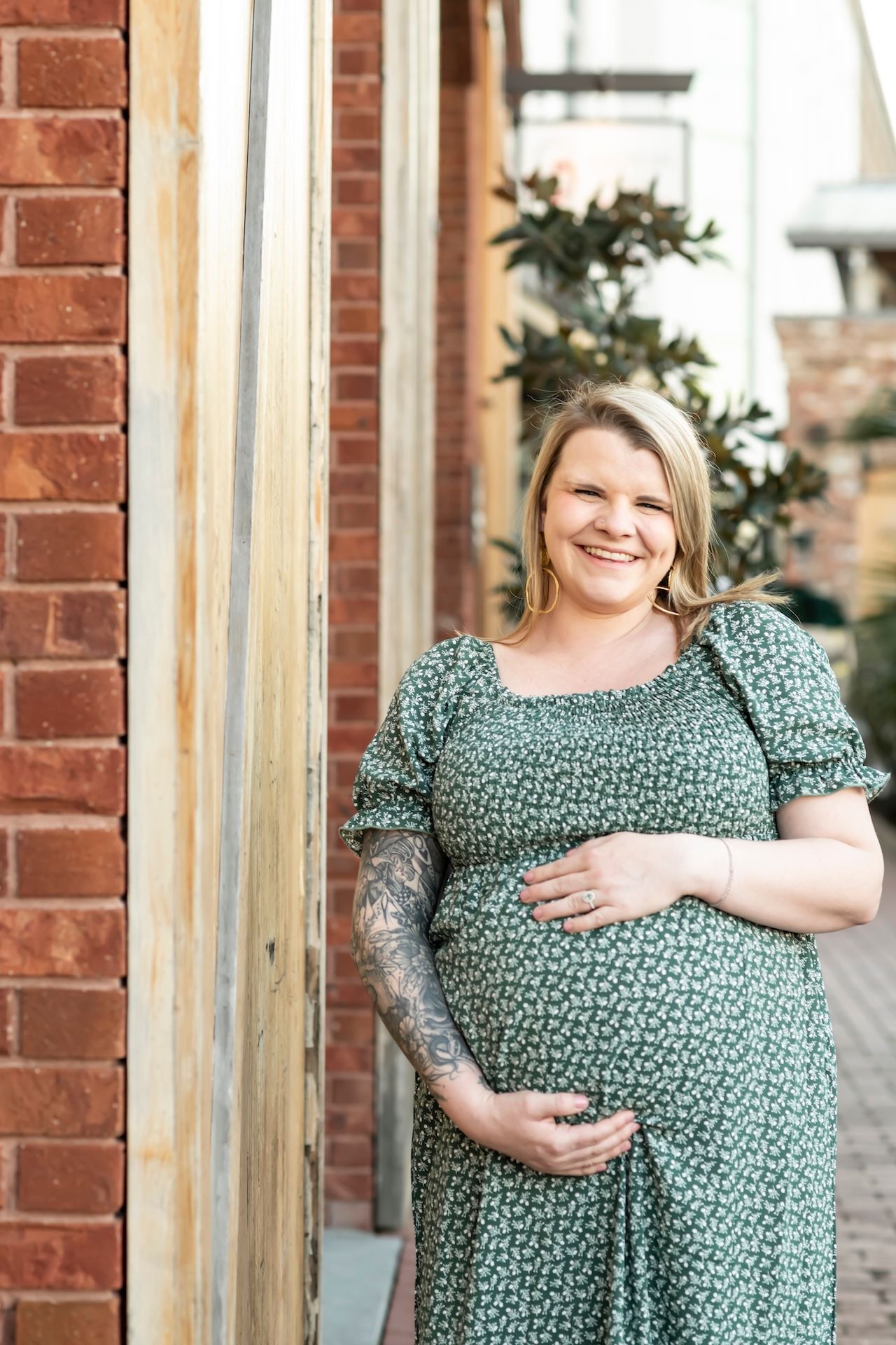 A person in a green floral dress stands against a brick wall while gently cradling their pregnant belly.