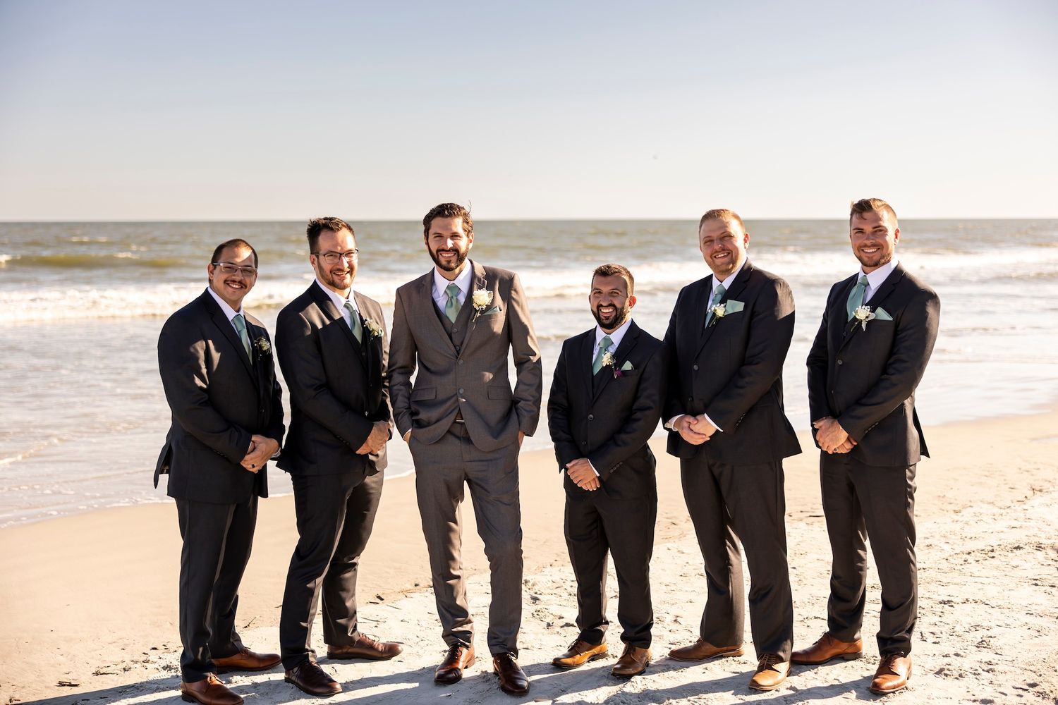 A group of groomsmen in black and gray suits stand together smiling on a sunny beach with ocean waves behind them.