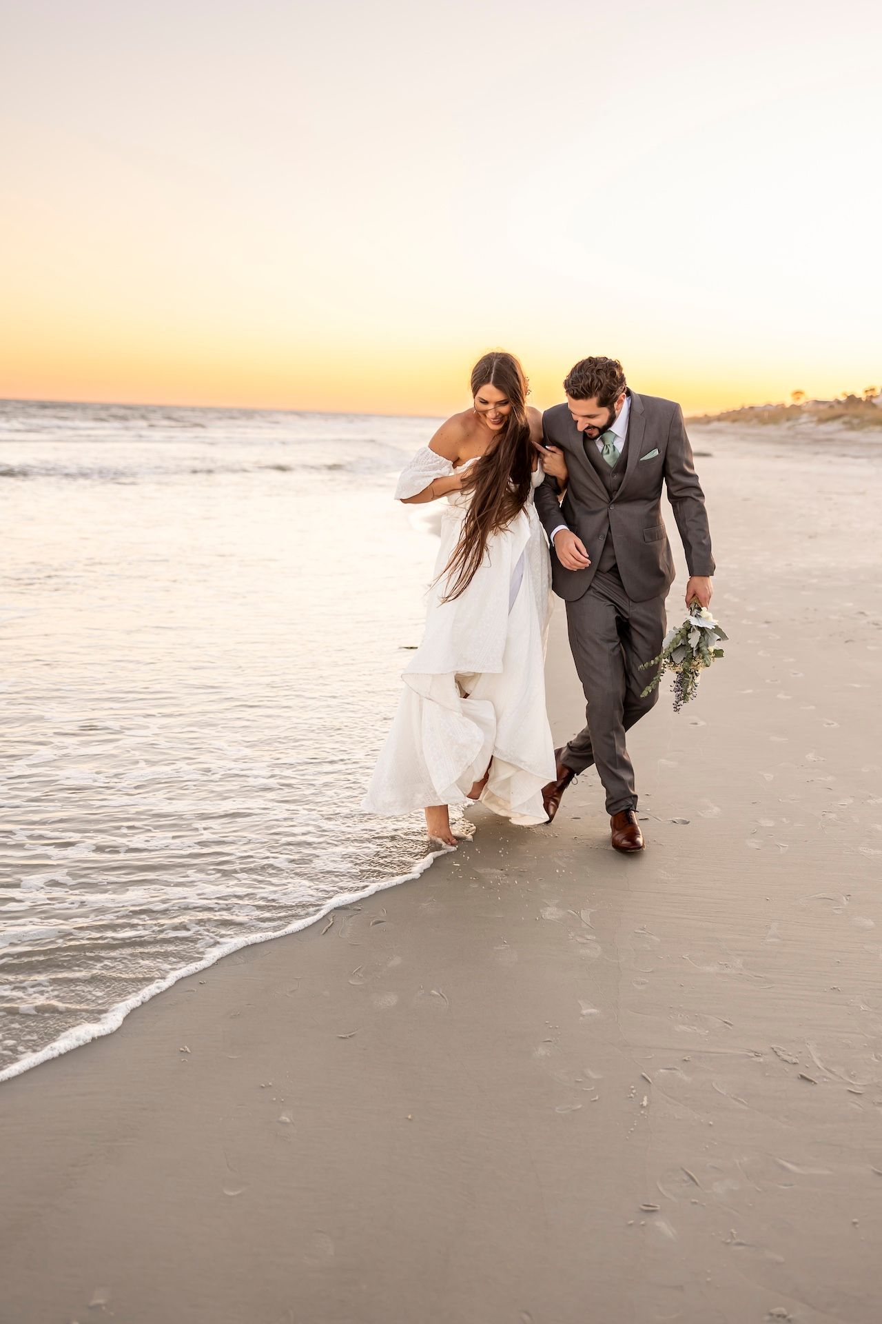 A couple in wedding attire walking together along a beach shoreline during golden hour sunset.
