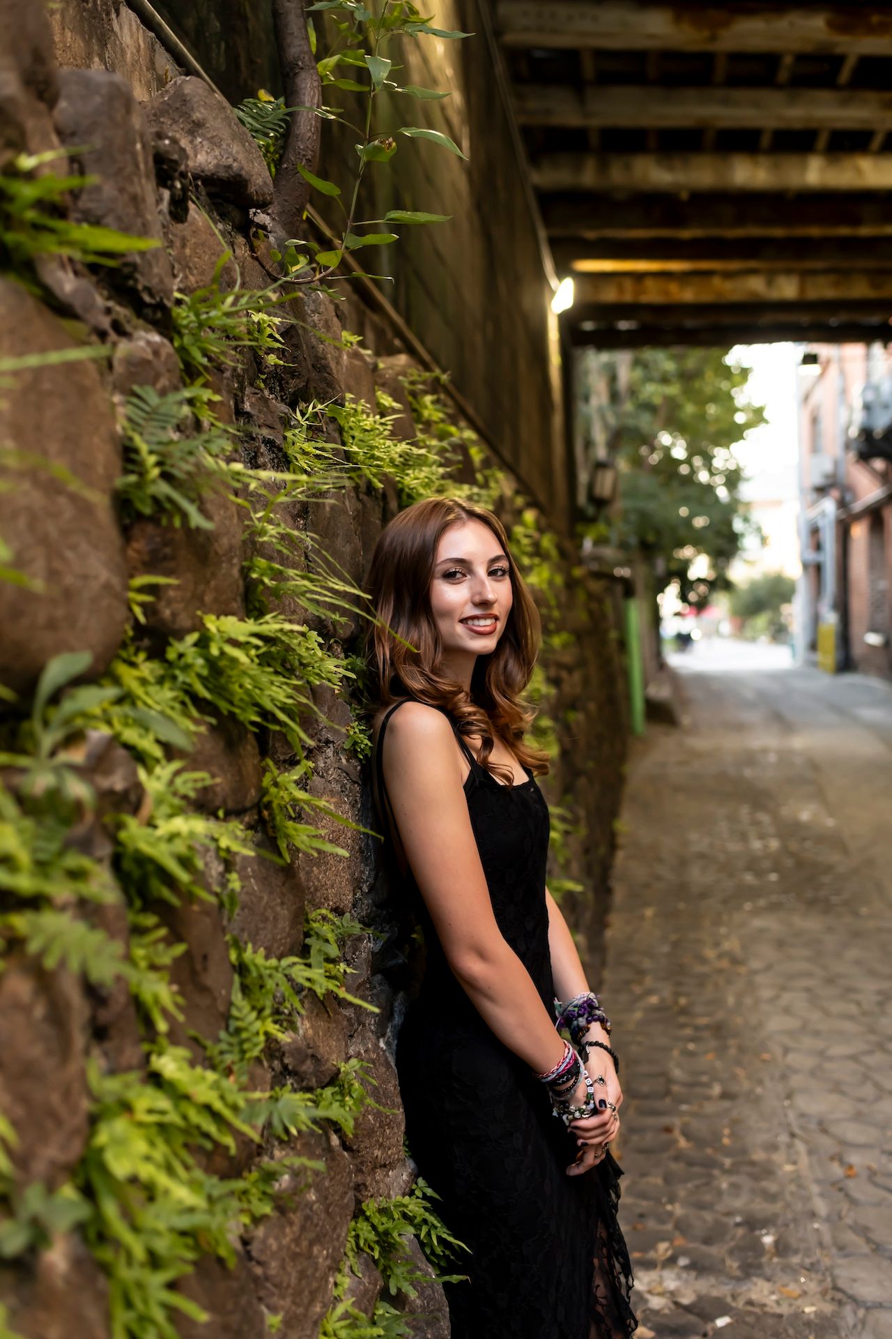 A black dress-clad figure stands against a mossy stone wall along a narrow cobblestone alley.