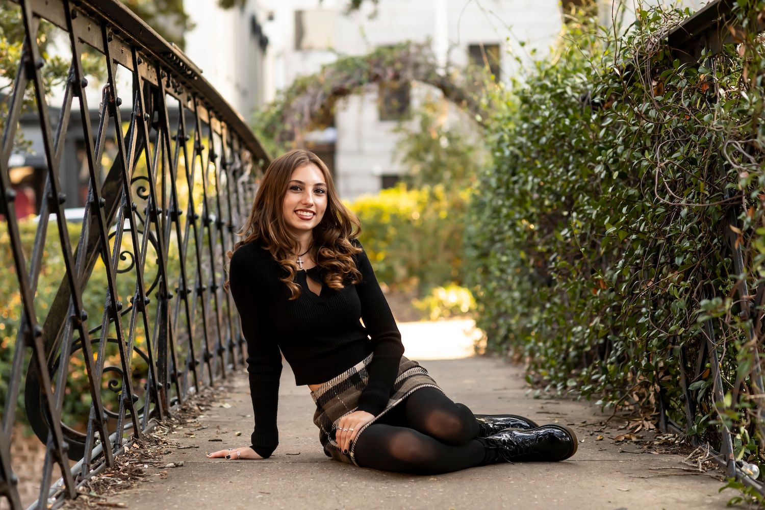 Person in black sweater and skirt sitting by ornate metal railing surrounded by lush garden foliage.